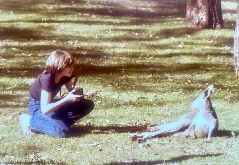 A woman kneeling on grass taking a photo of a kangaroo lying on the ground in a wooded park.
