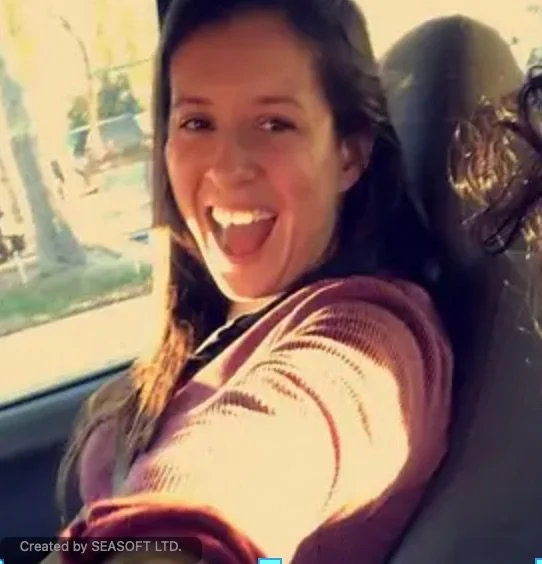 A young woman with brown hair taking a selfie in a car, smiling and showing her teeth, with sunlight coming through the window.
