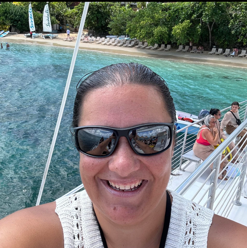 A woman smiling on a boat with sunglasses, with clear water and a sandy beach with lounge chairs in the background.