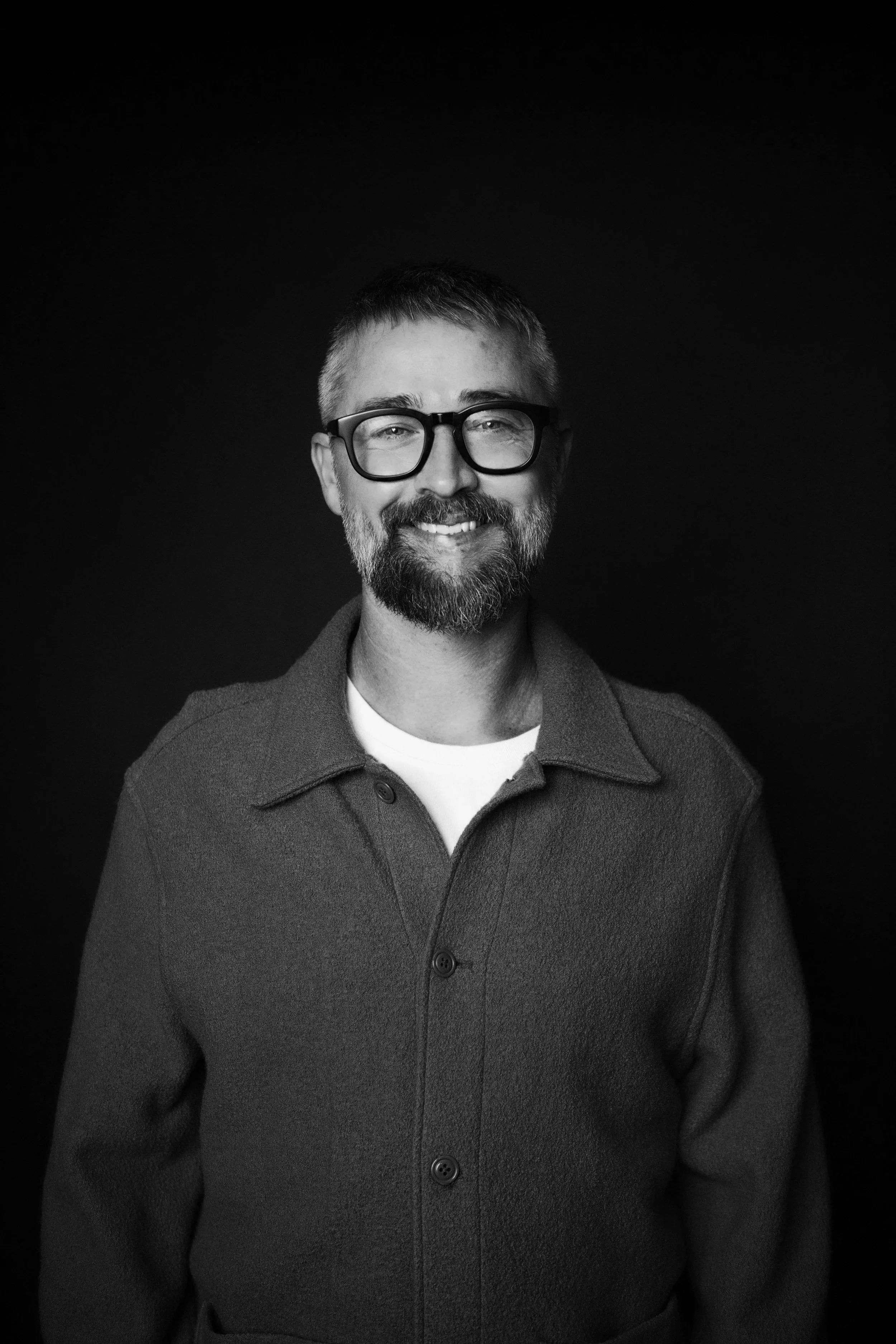 A black and white photo of a man with glasses, a beard, and short hair, smiling, wearing a coat over a shirt, against a dark background.