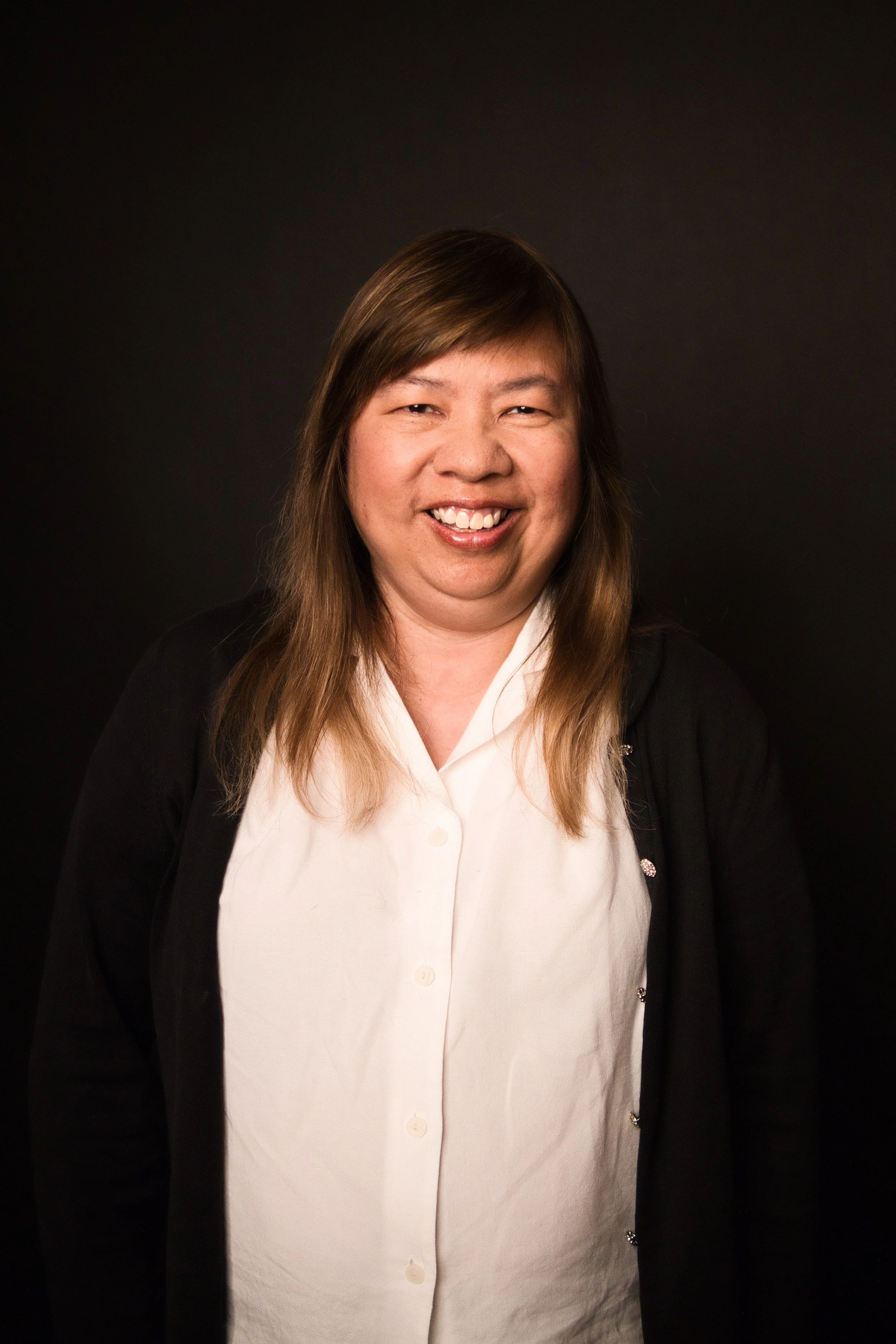 A woman with long brown hair, smiling, wearing a white button-up shirt and a black cardigan, standing against a dark background.