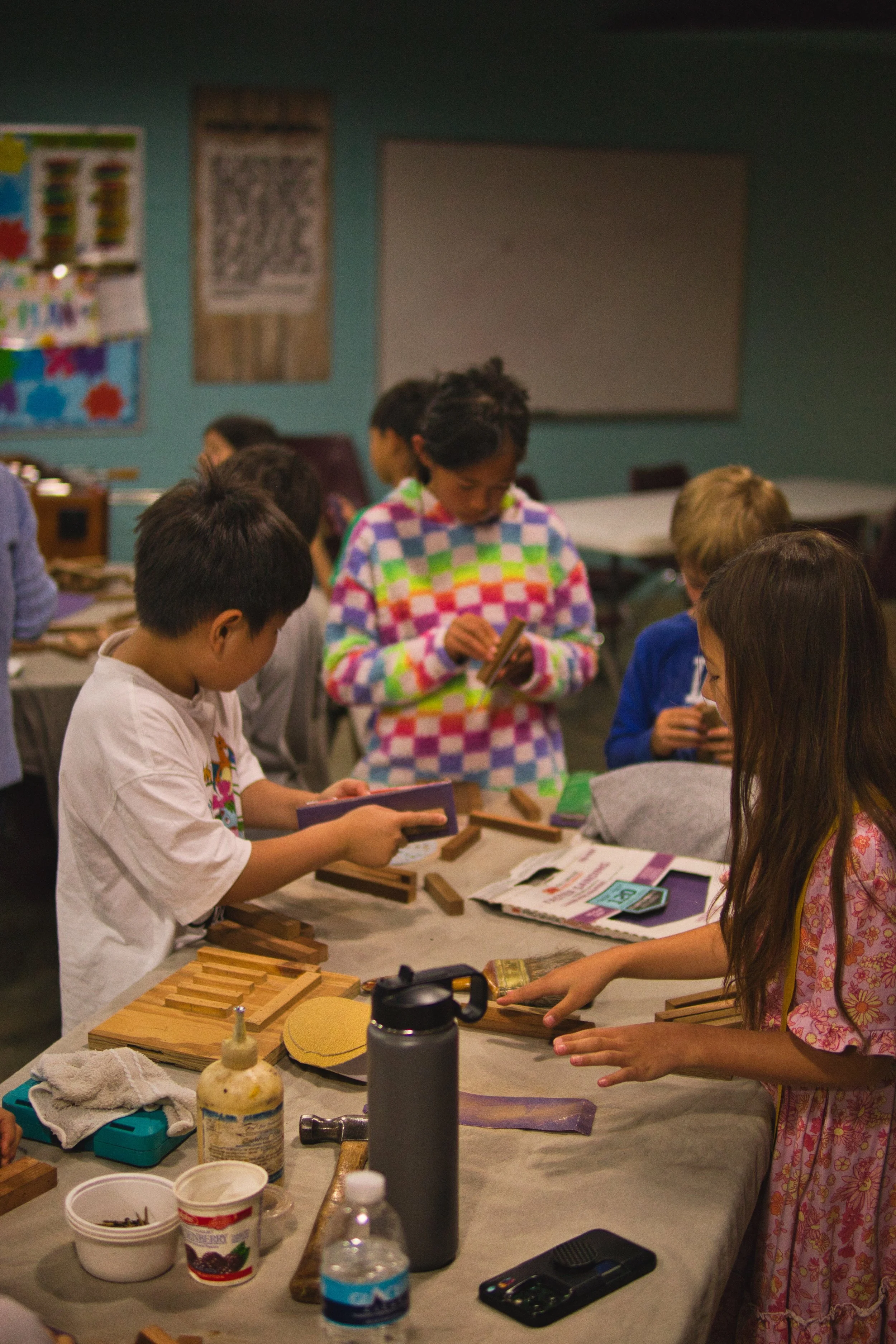 Children and adults gathered around a table engaging in a woodworking activity, working with wooden pieces and tools in a classroom setting.