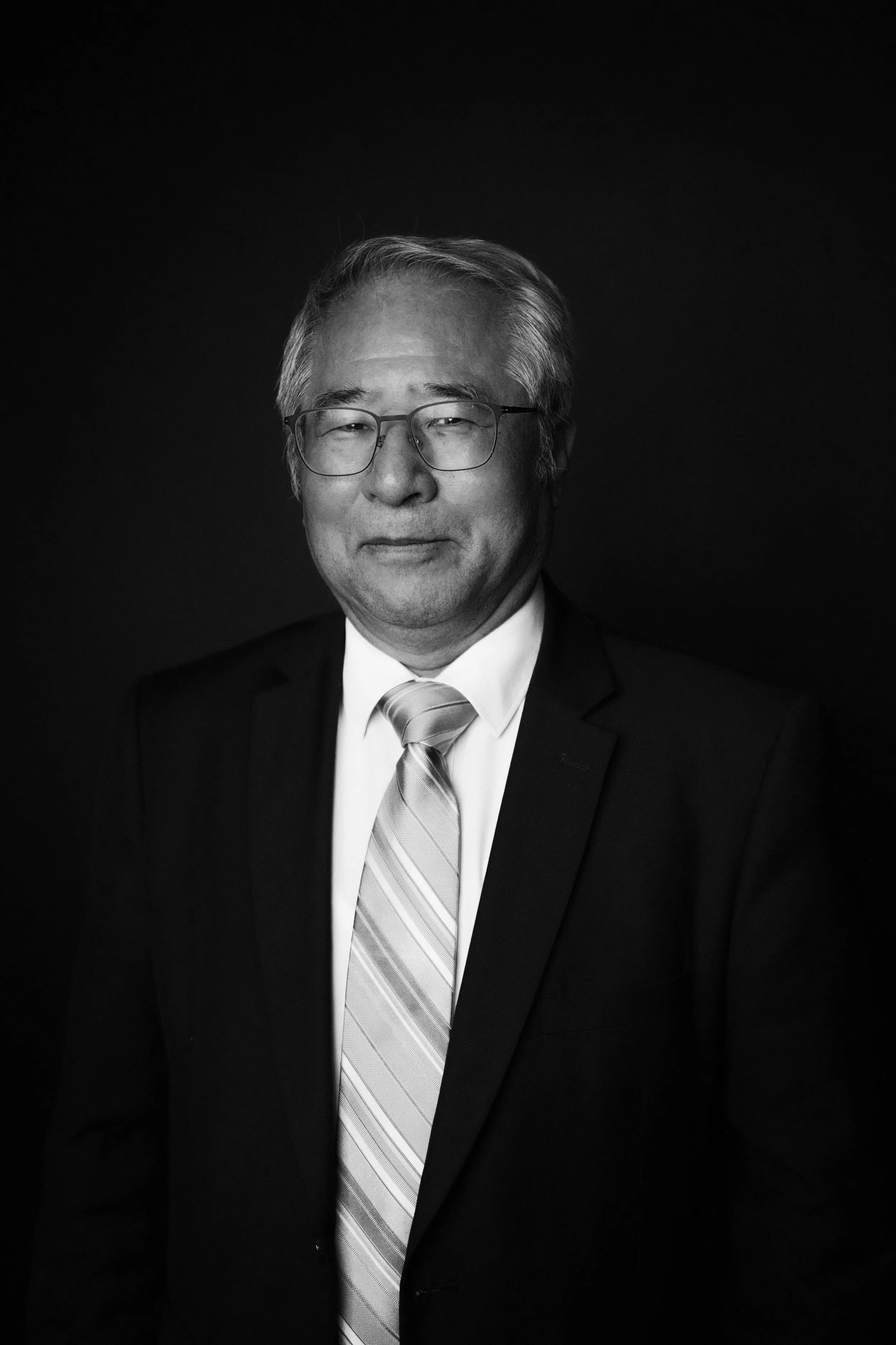 A black and white portrait of an older man wearing glasses, a suit, and a striped tie, smiling gently with a dark background.