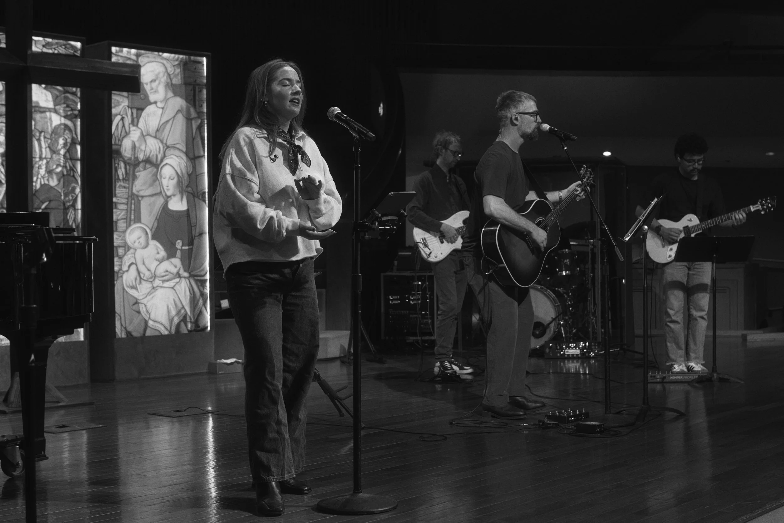 A group of four musicians performing on a stage, with two women and two men. The woman on the left is speaking into a microphone. The man on the right is playing an acoustic guitar. The stage has religious stained glass windows in the background.