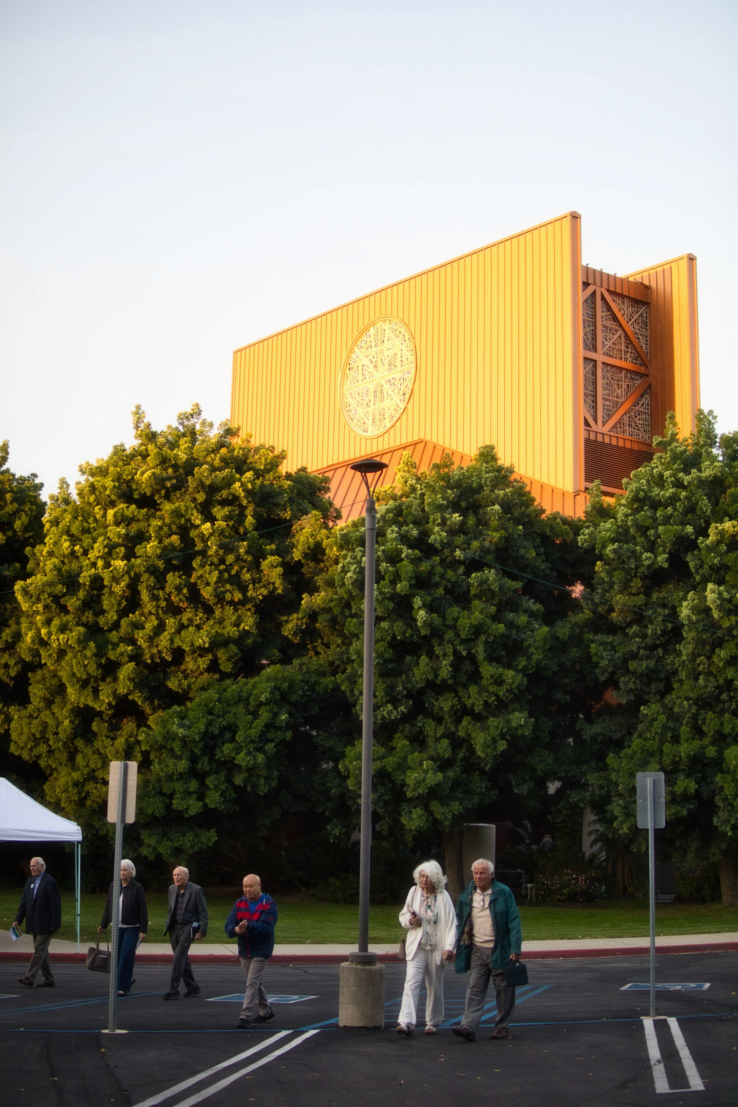 People walking in a parking lot in front of green trees with a large yellow building featuring a circular window and intricate design.