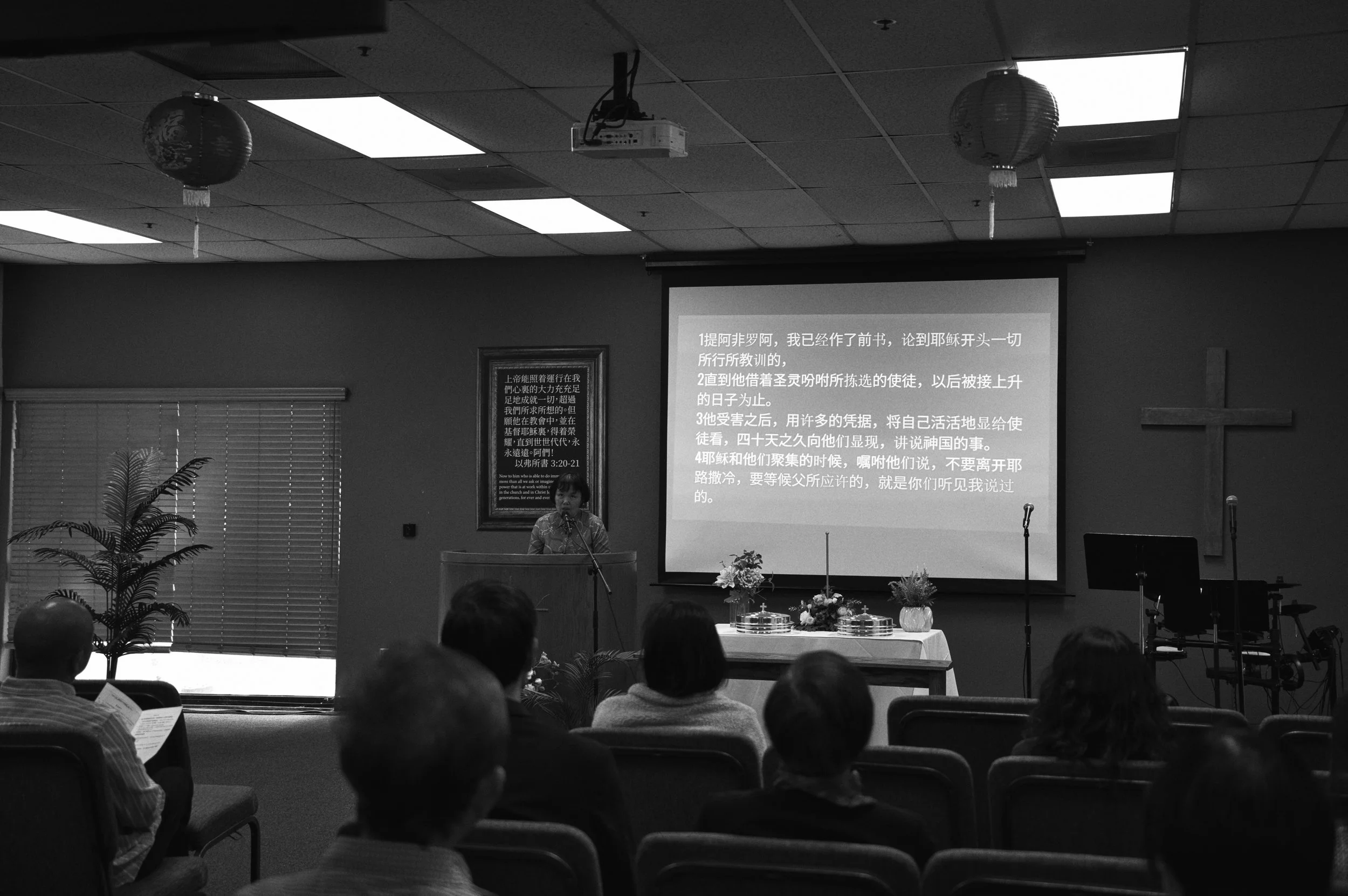 A woman standing at a pulpit speaking to an audience in a room decorated with plants, with a large screen displaying Chinese text and a framed sign on the wall.