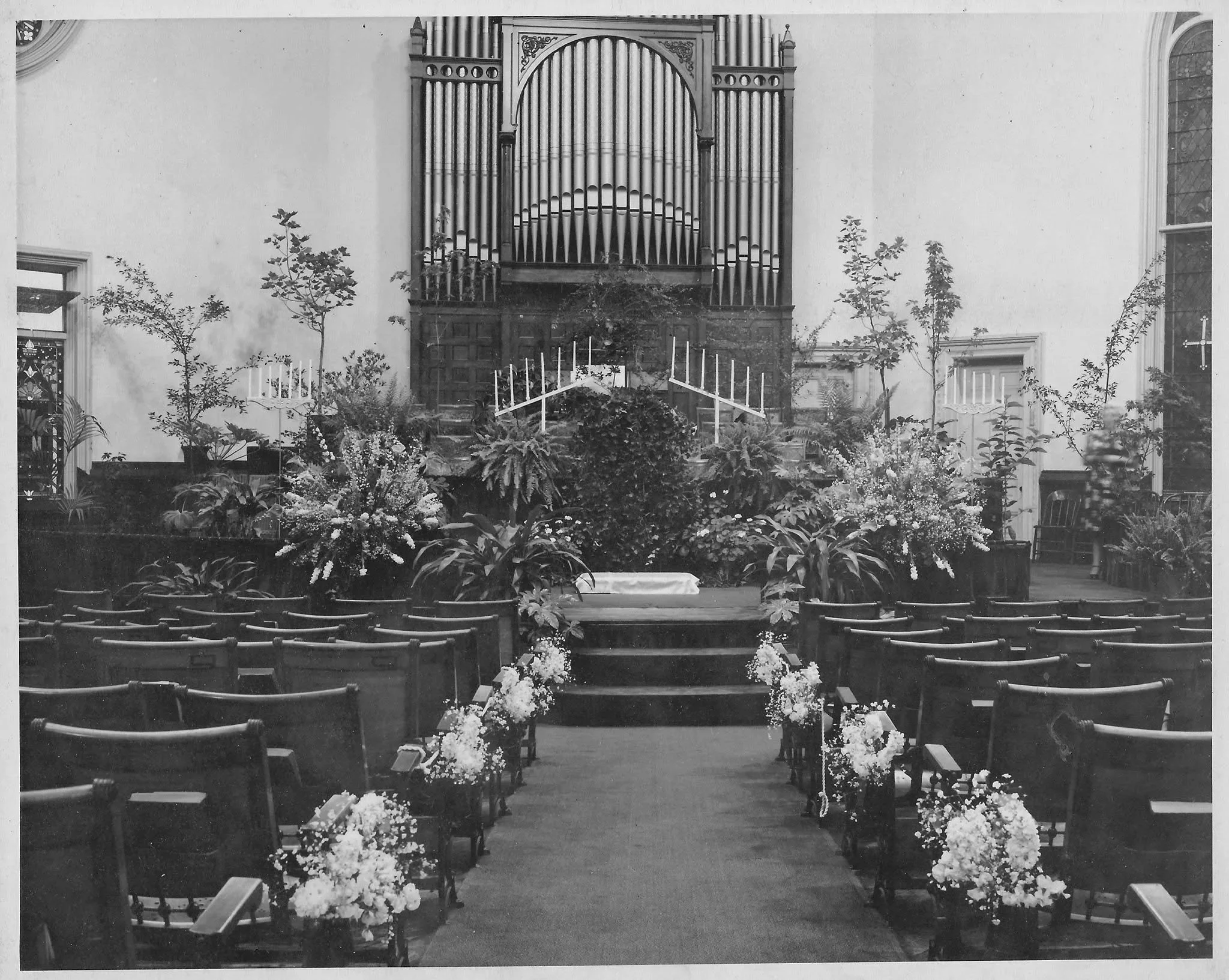A black-and-white photo of the interior of a church with an altar decorated with flowers, with a large pipe organ in the background, and pews arranged in rows facing the altar.