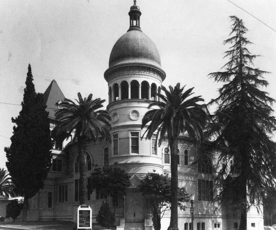 A historic building with a large dome, surrounded by palm trees and other tall trees, under a cloudy sky.