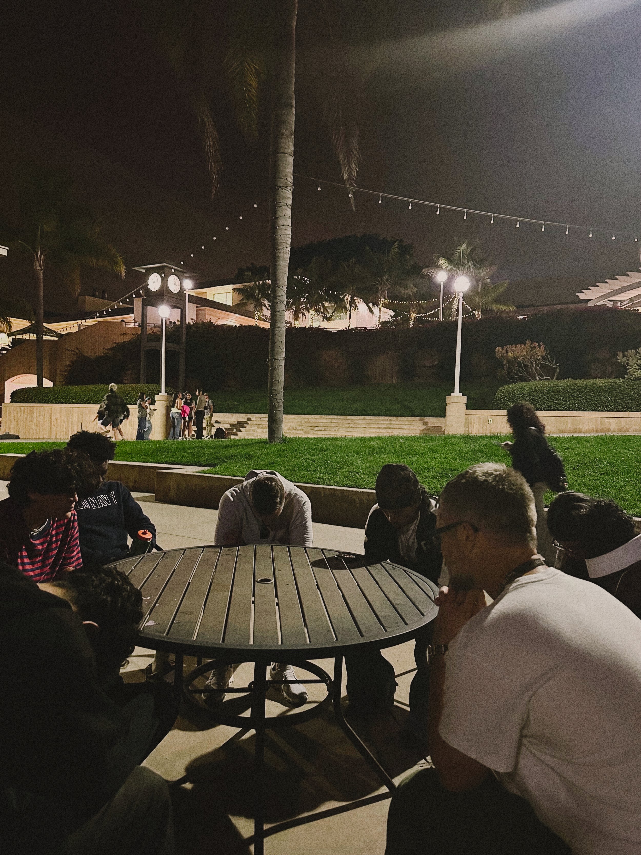 People sitting and socializing around a round table in a park at night, with illuminated trees, streetlights, and a clock tower in the background.