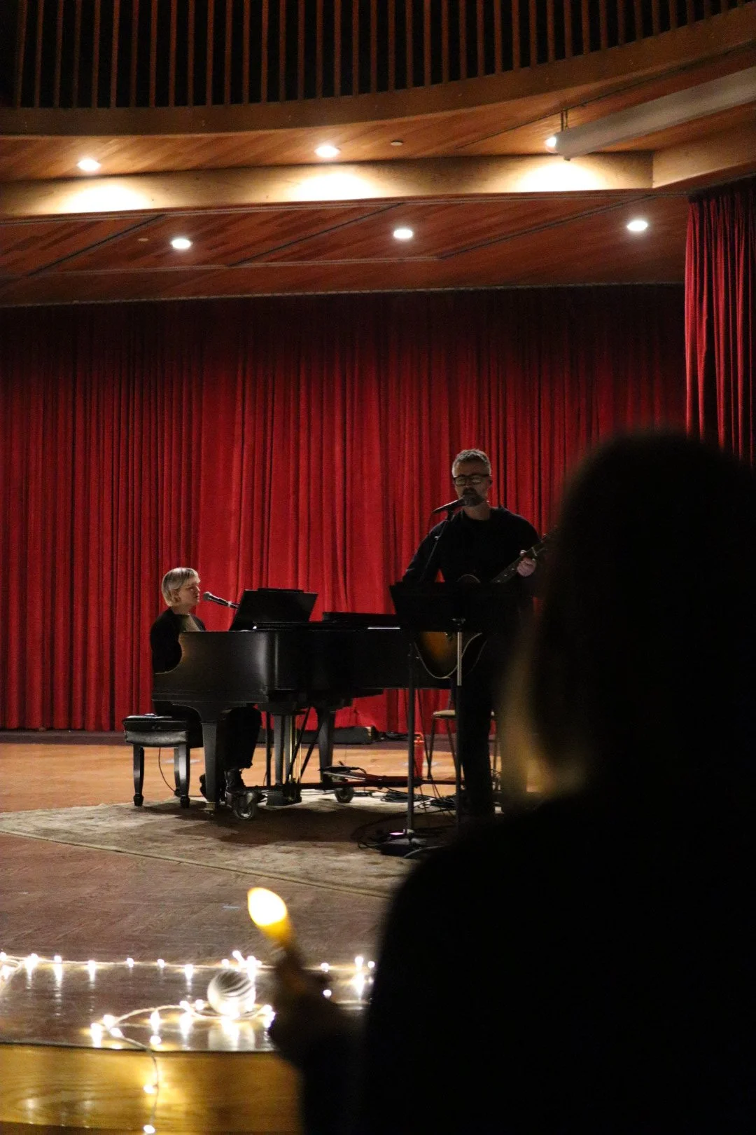 Musicians performing on stage with a woman playing the piano and a man singing and playing guitar, in front of red curtains, under warm lighting.