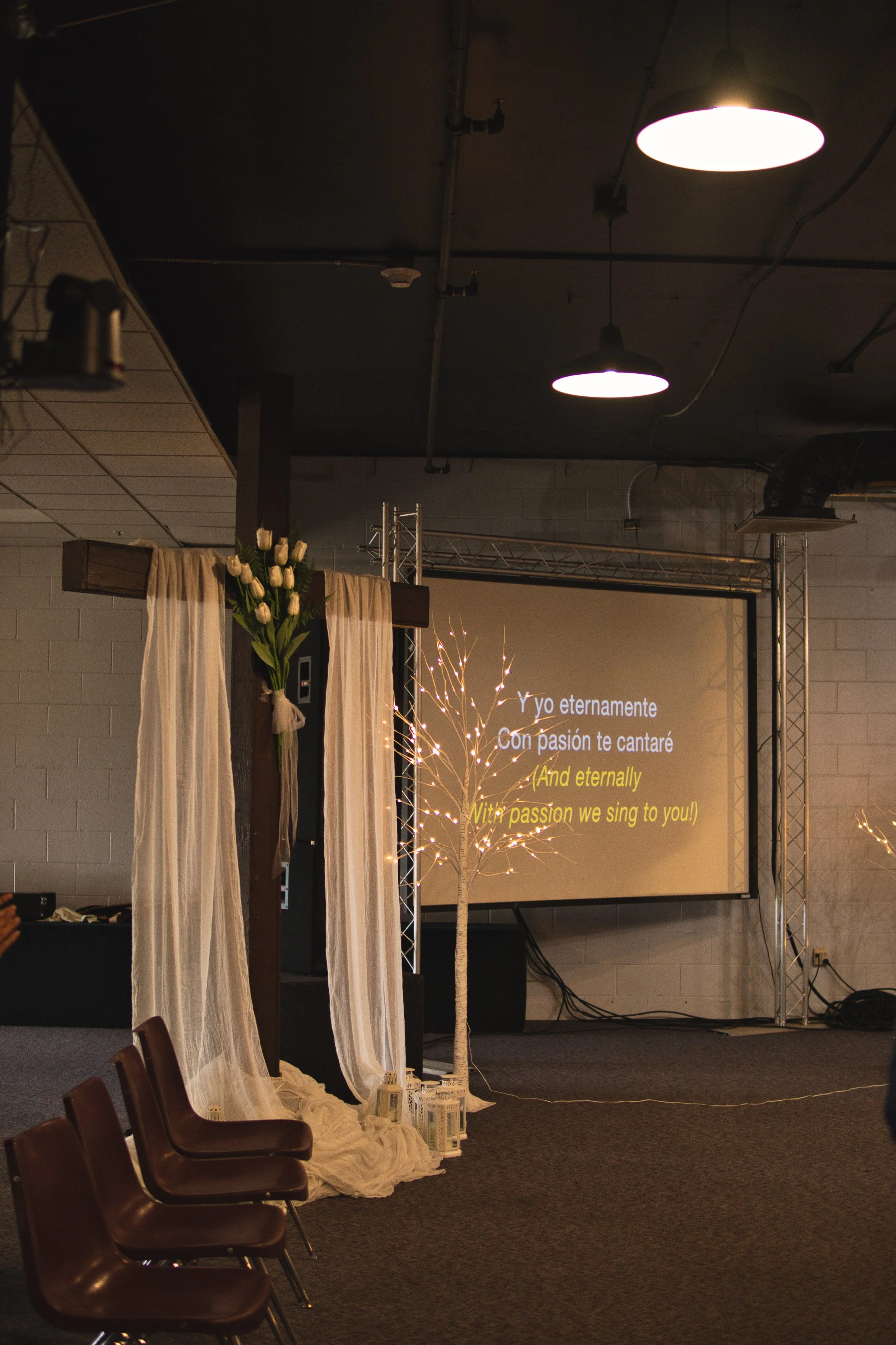 Indoor event space with chairs, decorative cross with white flowers, sheer curtains, illuminated tree, and a projected screen displaying song lyrics in Spanish and English.
