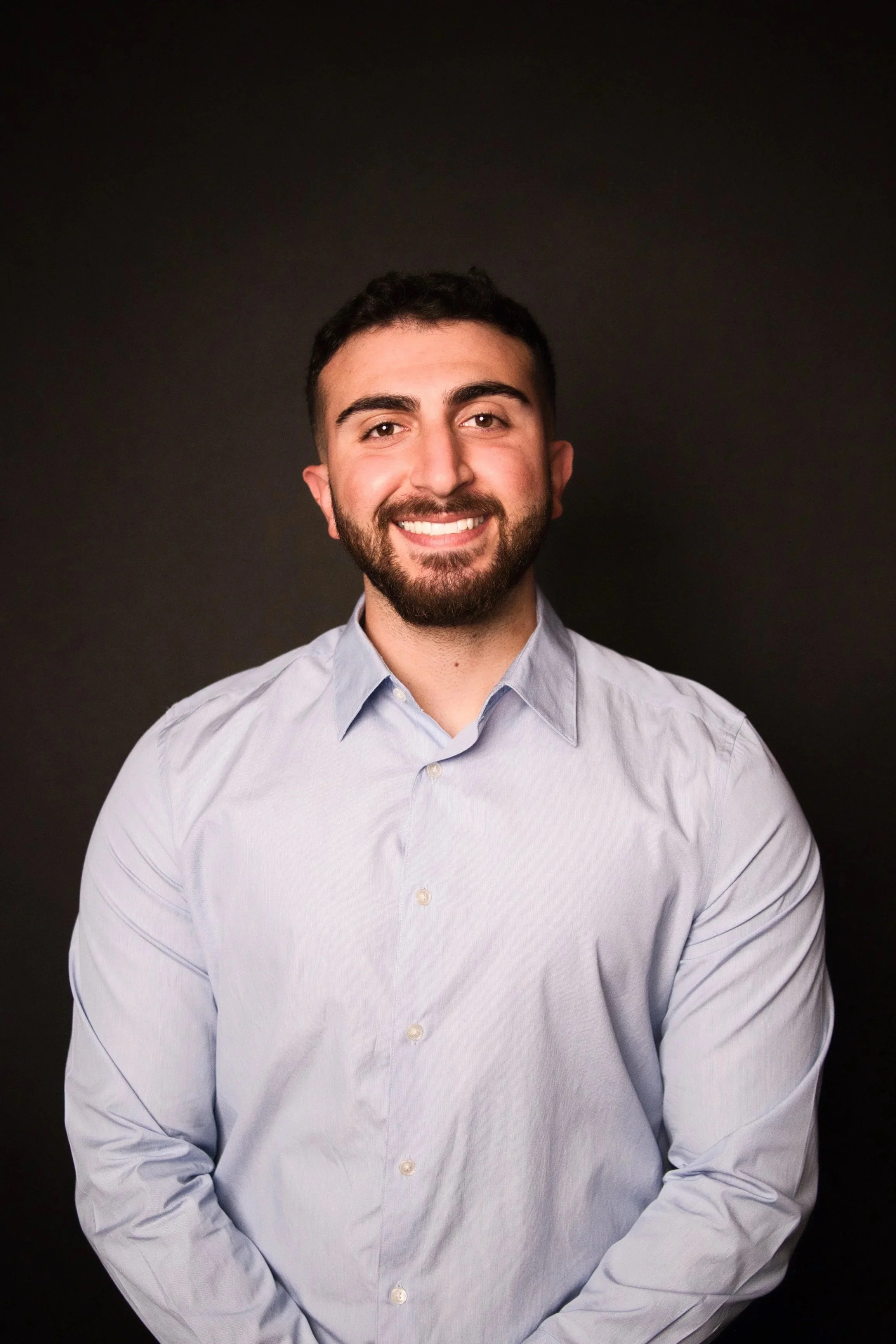 Portrait of a smiling young man with dark hair and a beard, wearing a light blue button-up shirt, standing against a black background.