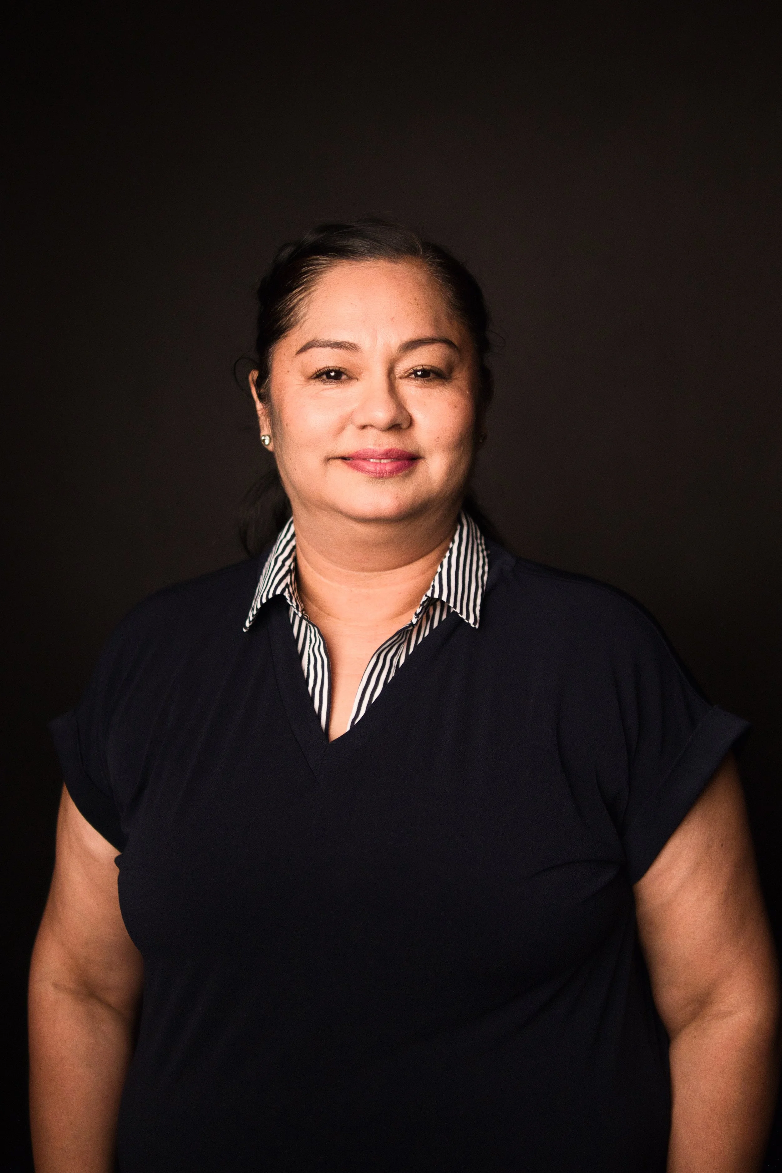 A woman with dark hair, wearing earrings, a black top, and a striped collared shirt underneath, standing against a plain dark background.