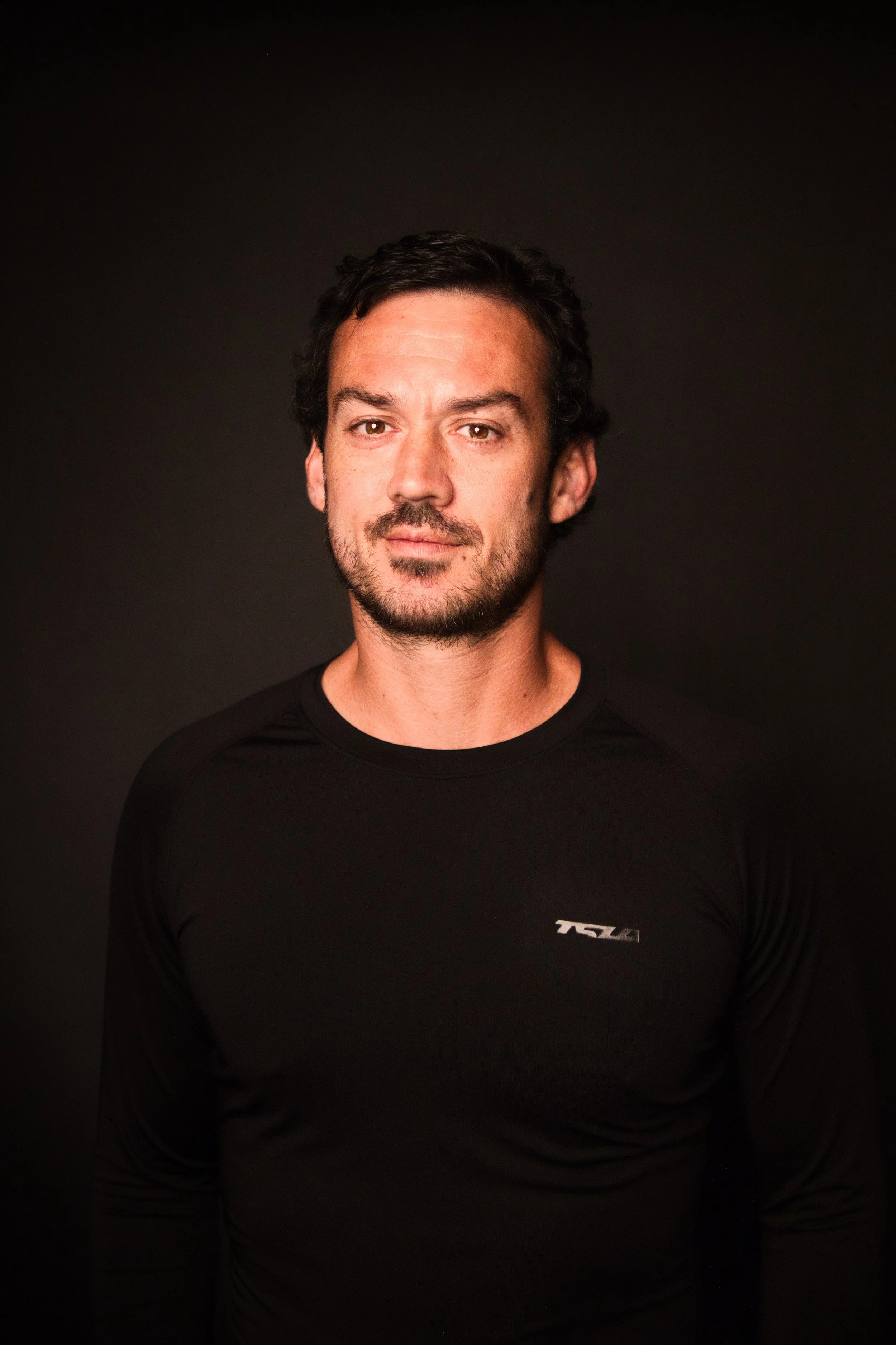 Portrait of a man with dark hair and beard, wearing a black long-sleeve shirt, against a black background.