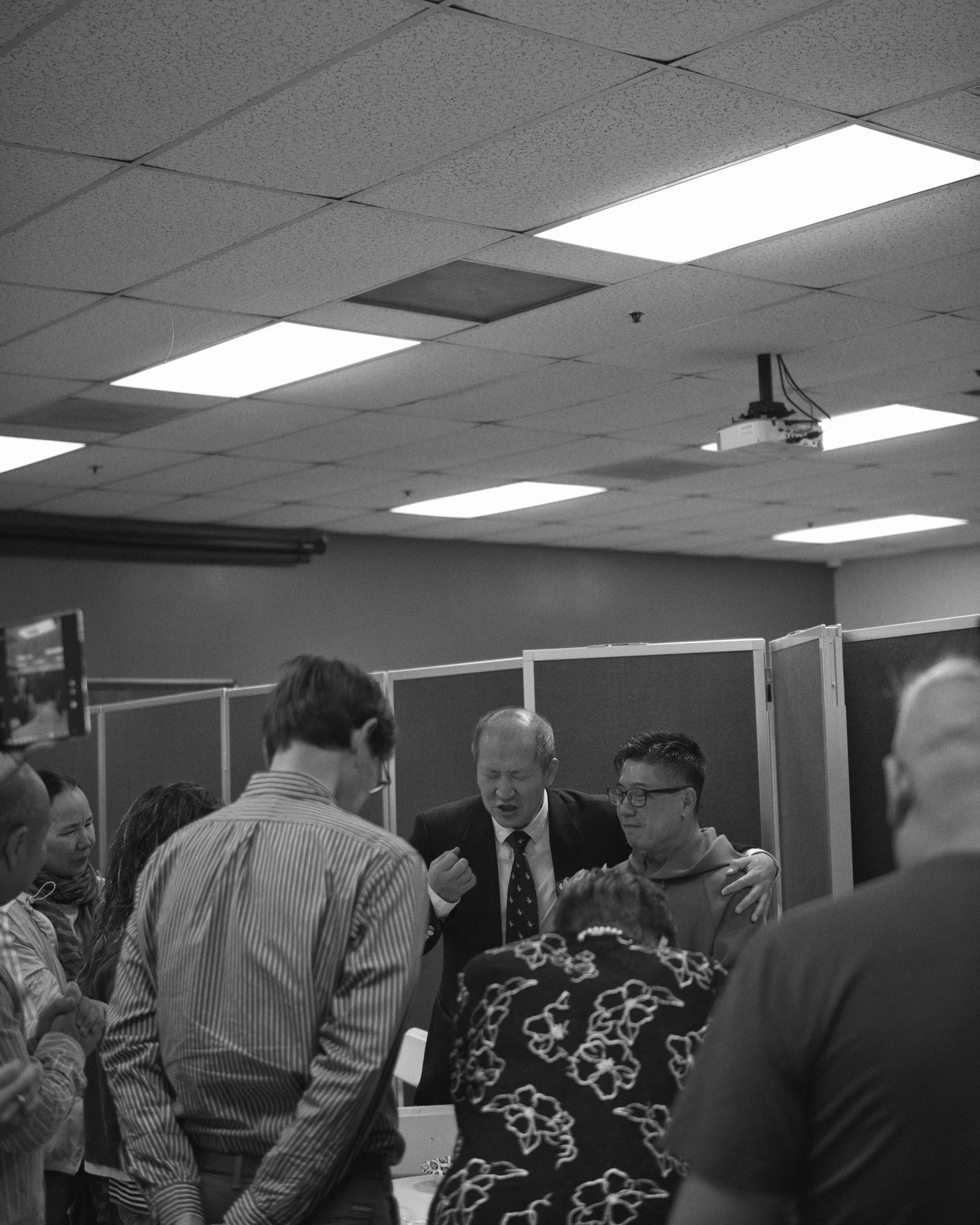 A group of people standing in a room, engaged in a discussion or prayer, with some individuals embracing or holding hands, and a ceiling with fluorescent lights and ceiling tiles.