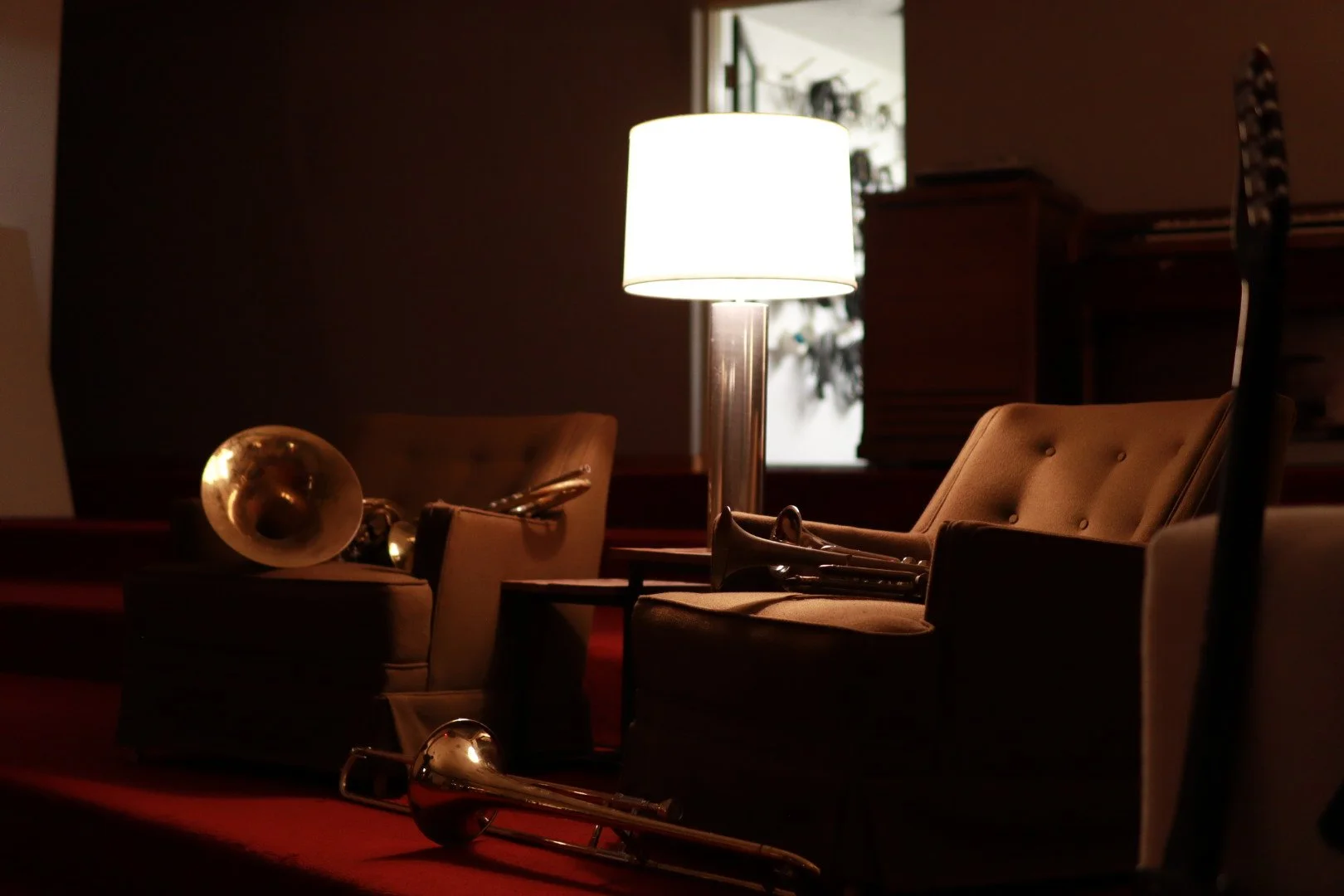 Interior of a cozy room with chairs, a table, a lamp, and musical instruments including a trumpet and a French horn, with warm lighting.
