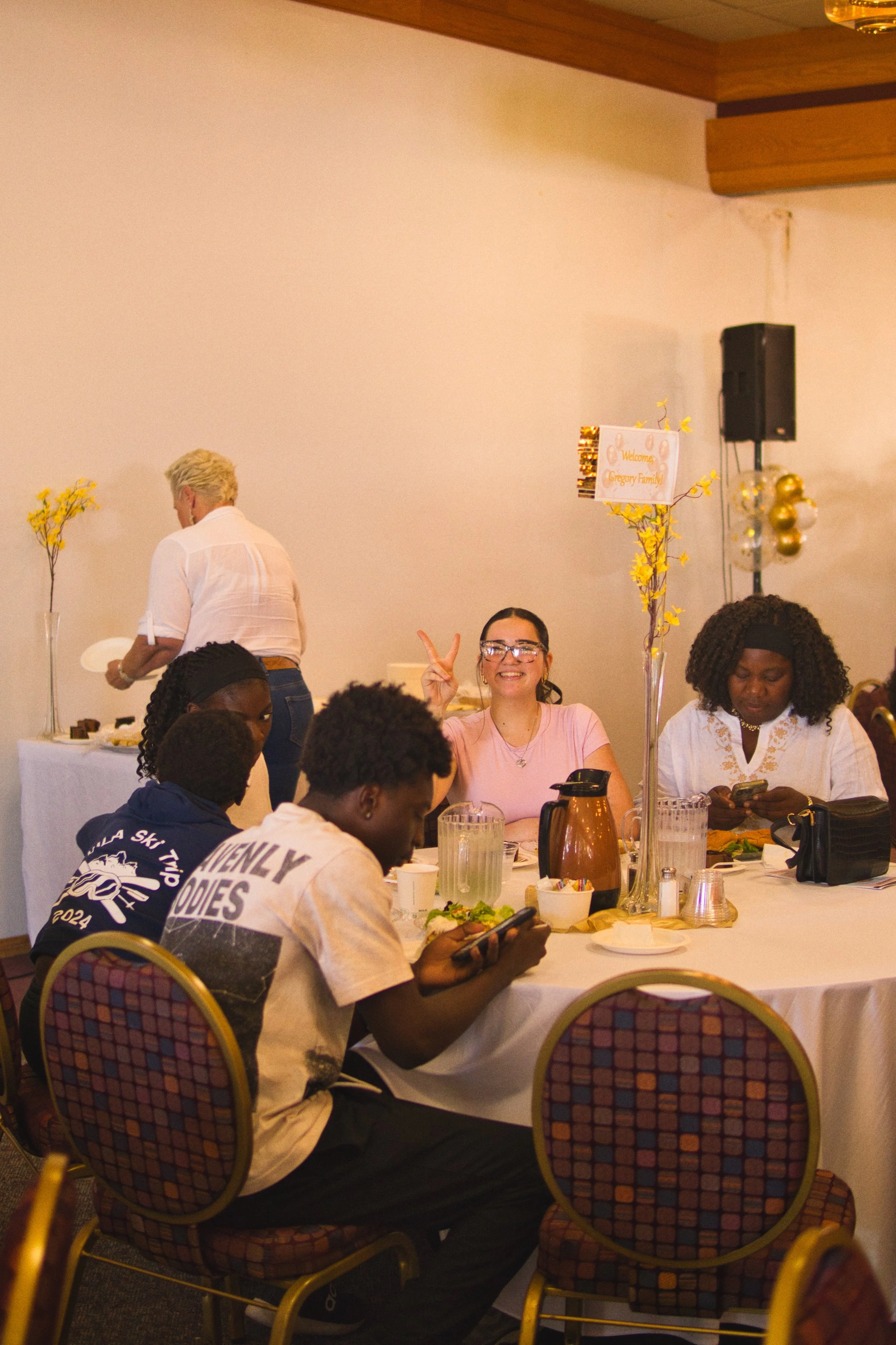 People sitting and standing around a decorated table at a gathering, with a large sign that reads 'Welcome Gregory Family', and a woman smiling and making a peace sign in the background.