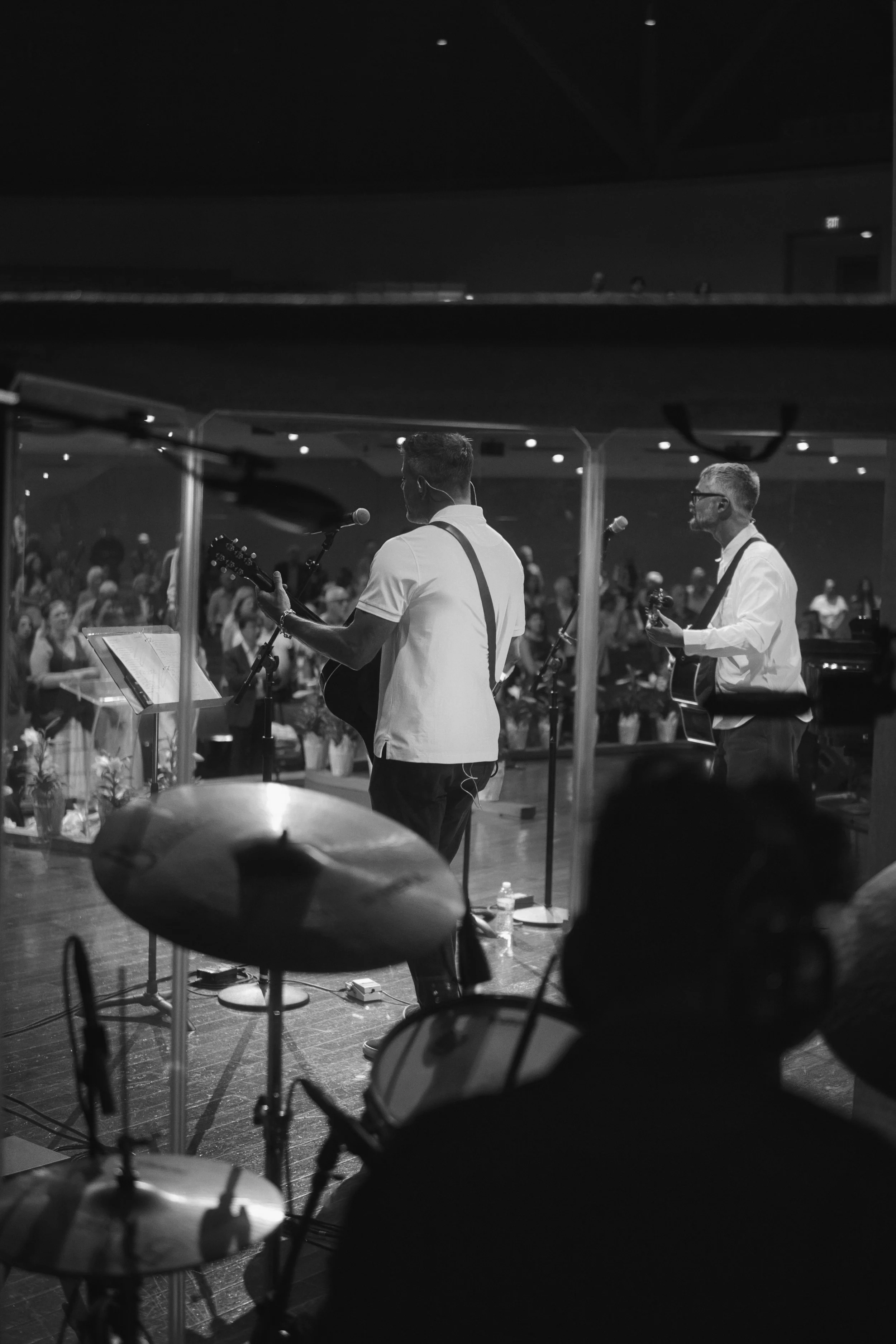 Black and white photo of a musical performance with two guitarists, one playing an acoustic guitar and the other an electric guitar, on stage in front of an audience. The photo shows drum cymbals in the foreground, and a crowd seated in the background.