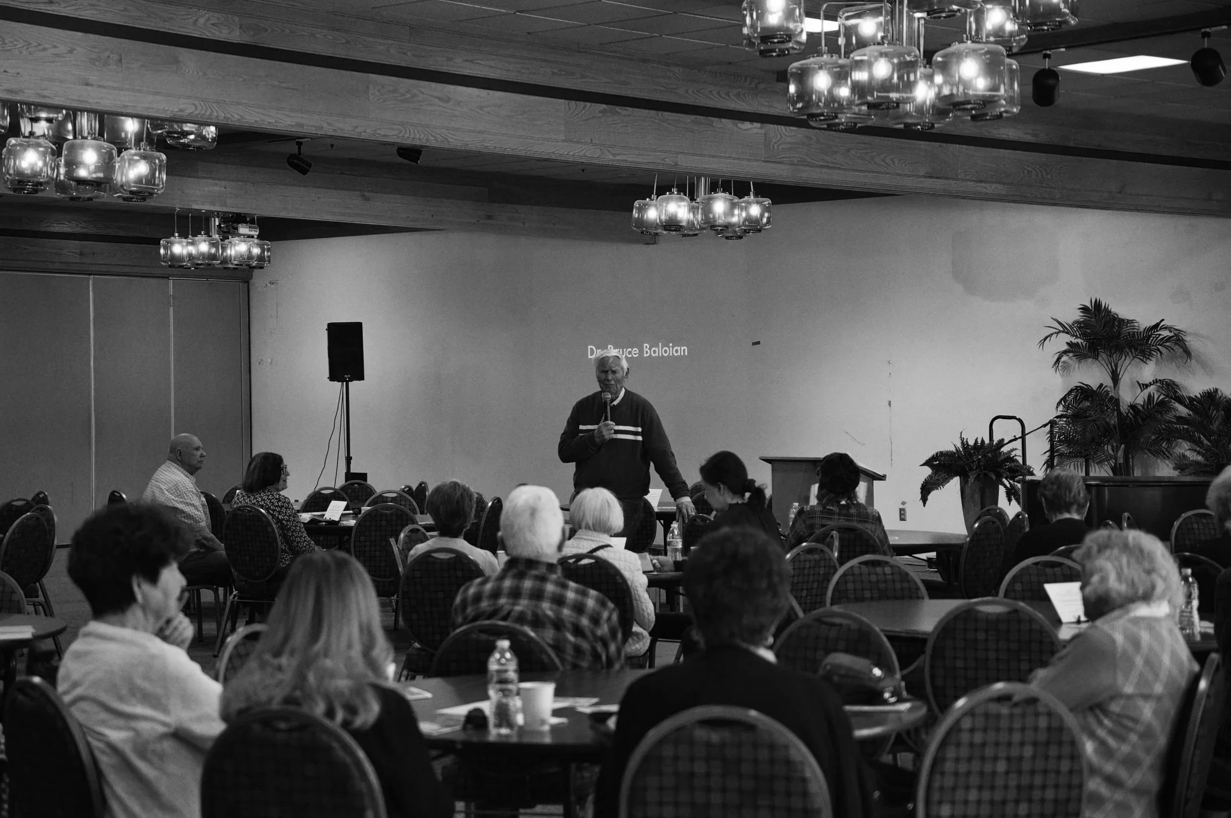 A man with gray hair speaking into a microphone during a presentation in a conference room filled with seated attendees. The room has wooden ceiling panels, modern hanging light fixtures, and potted plants.