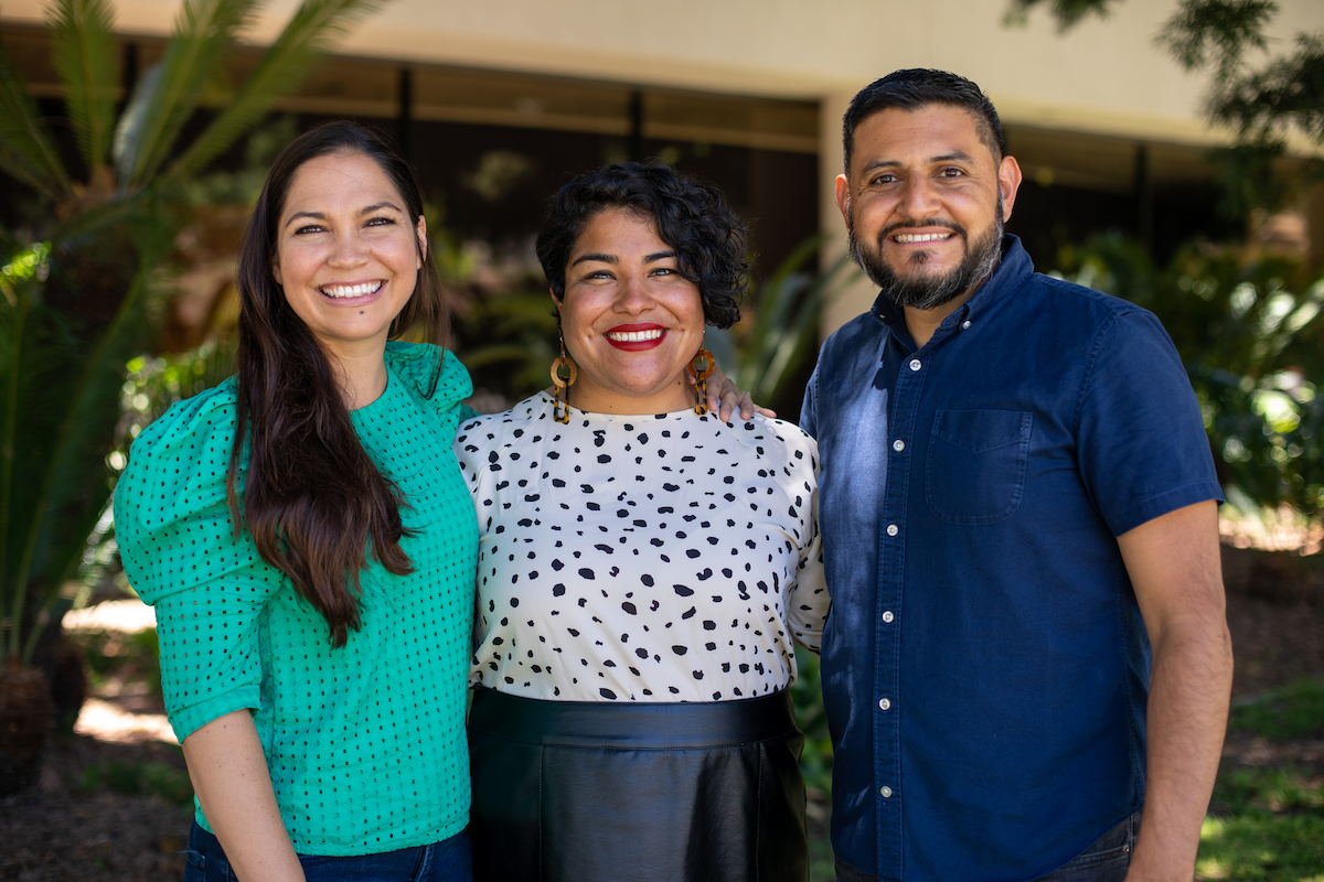 Three people standing outdoors, smiling at the camera, with trees and a building in the background.
