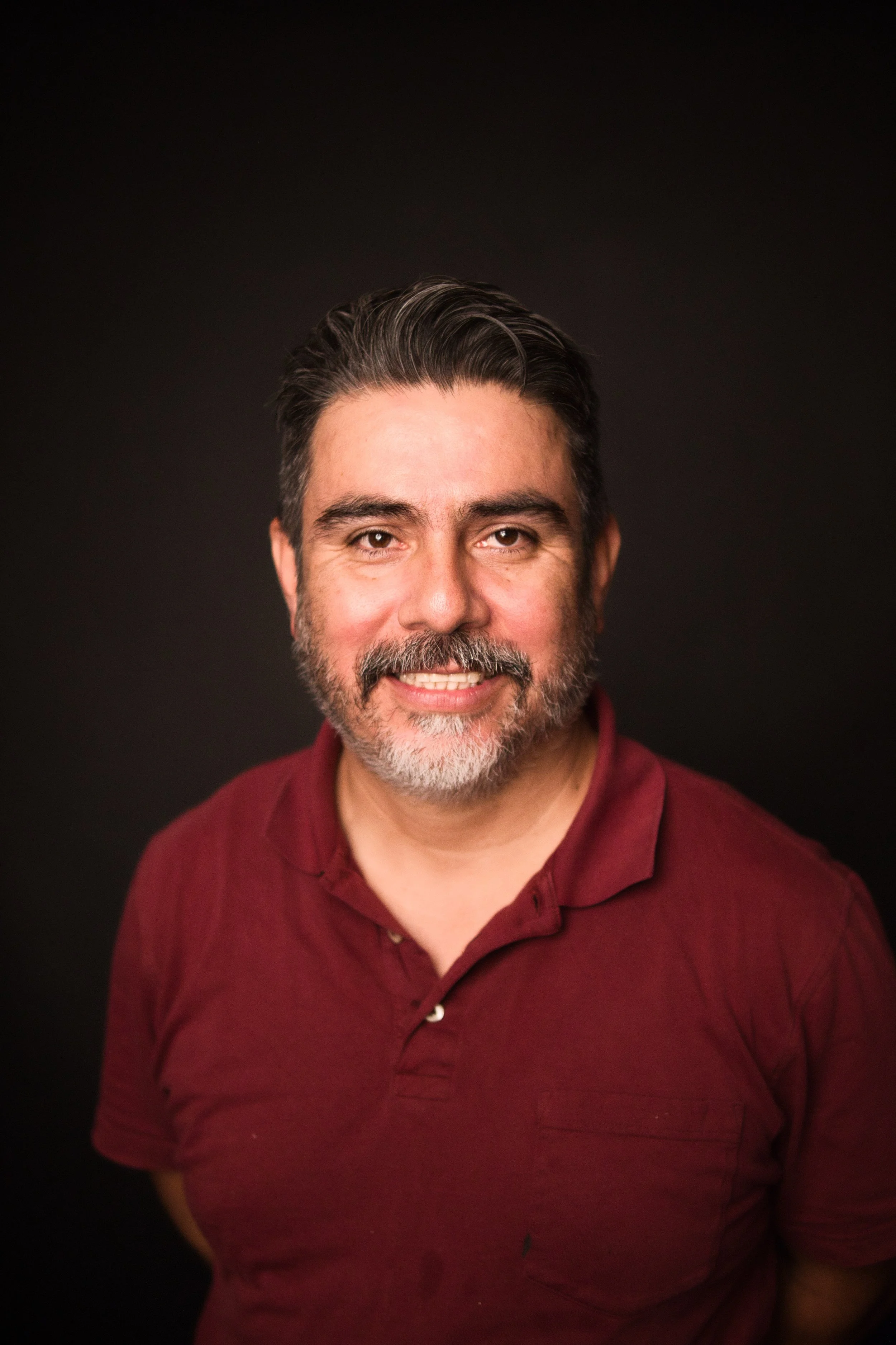 A middle-aged man with dark hair and a beard, smiling, wearing a maroon collared shirt against a dark background.