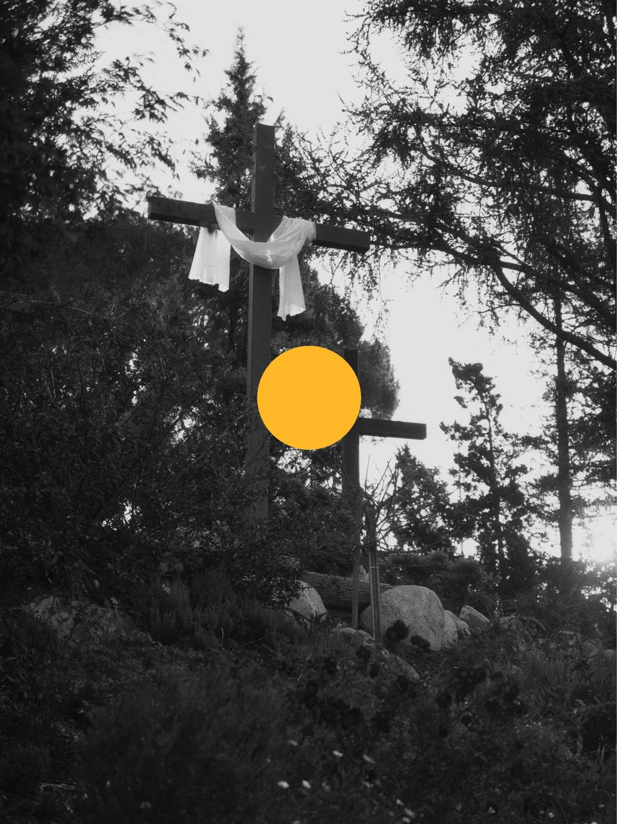 A black and white photograph of a wooden cross with white cloth draped over it, set outdoors among trees and rocks, with a large yellow circle partially obscuring the lower part of the cross.