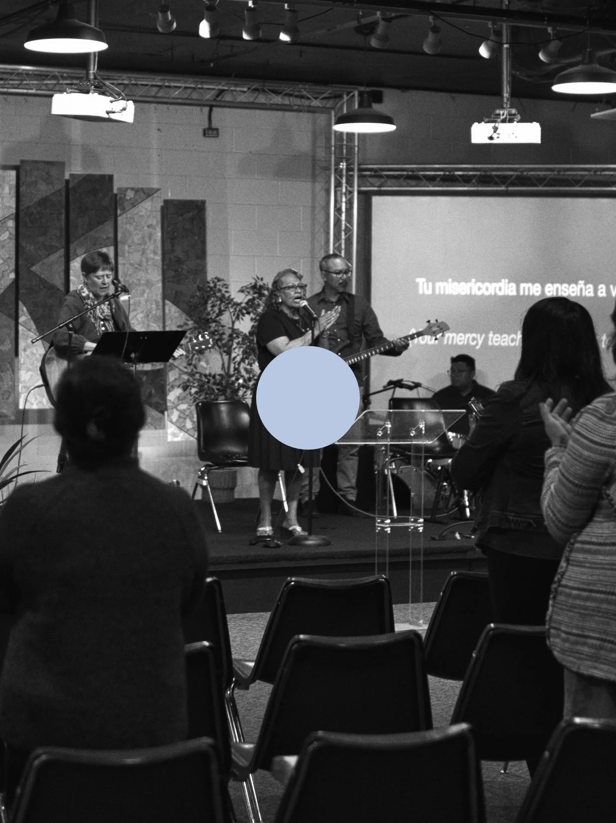 A church service or worship event with musicians on stage, including a woman singing, a man playing guitar, and another woman playing guitar, with people in the audience clapping and listening, a large screen displaying lyrics in Spanish and English, and stage lighting above.