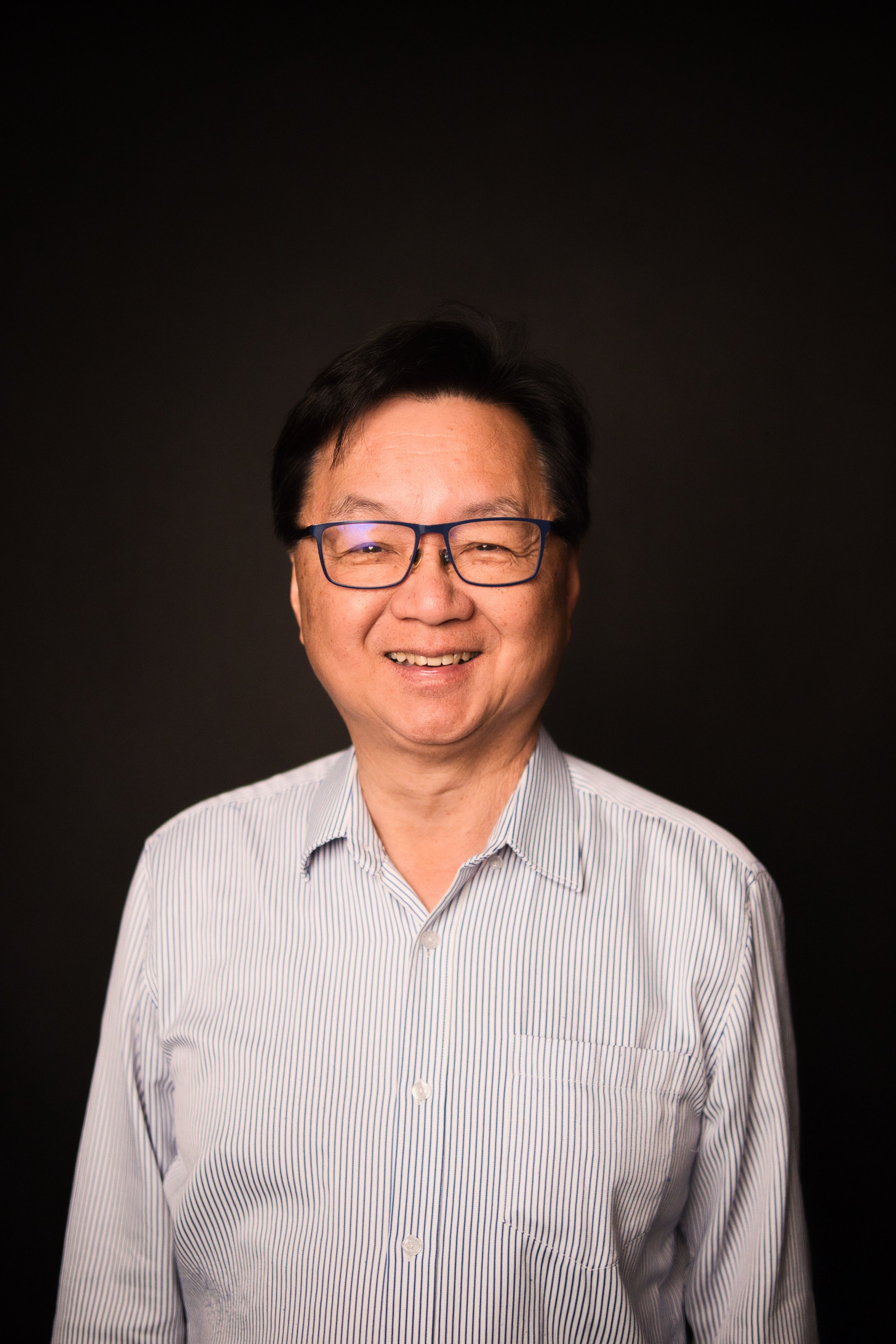 A man with glasses and dark hair smiling, wearing a light-colored striped button-up shirt, against a black background.