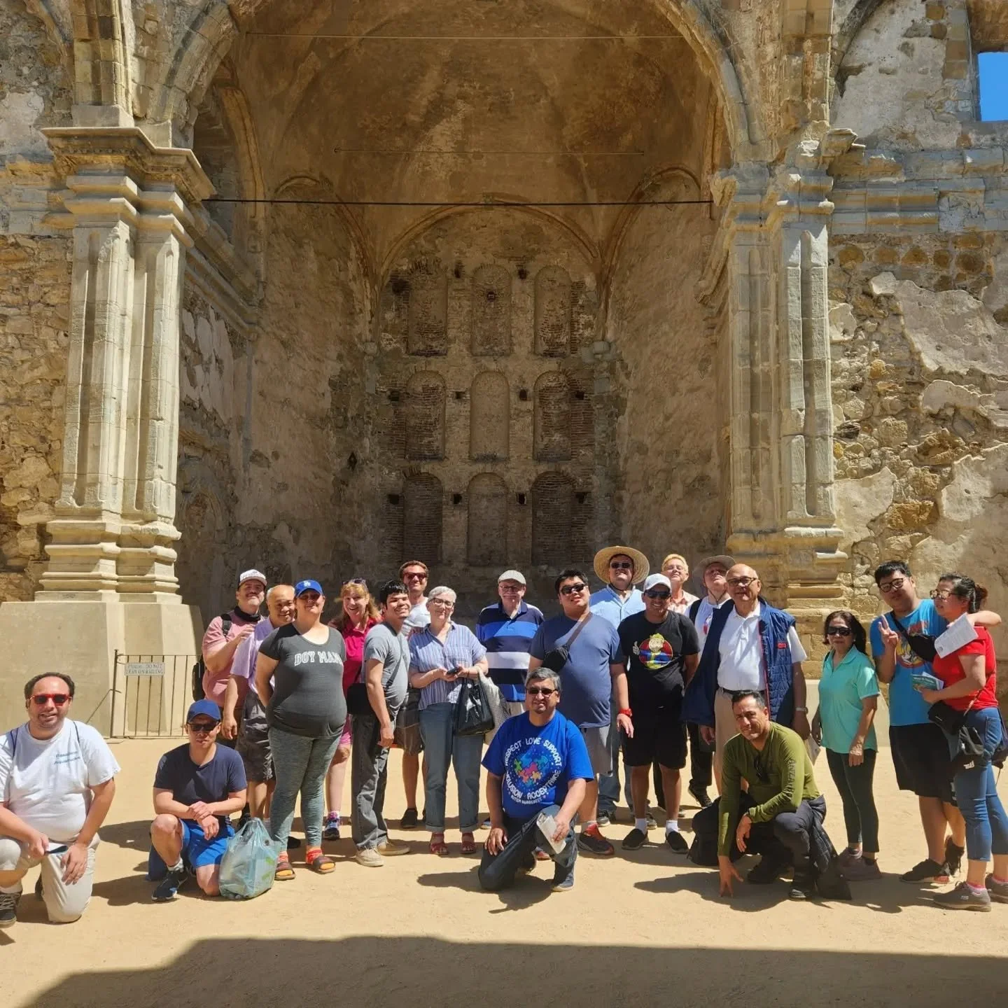 Group of tourists posing in front of ancient ruins with stone walls and arches, some wearing hats and sunglasses, under a clear blue sky.