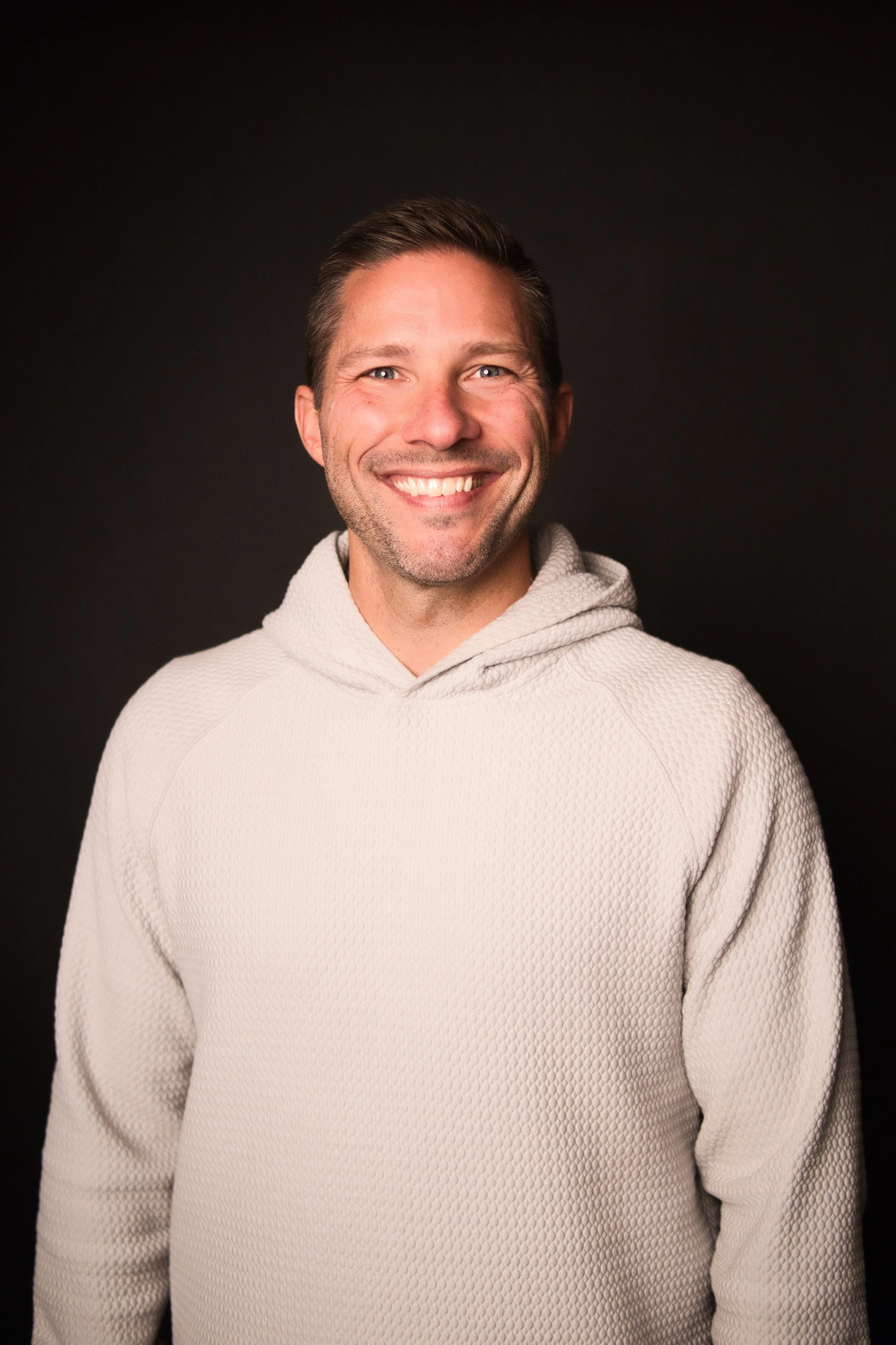 A smiling man with short brown hair wearing a light-colored, textured hoodie, against a plain black background.