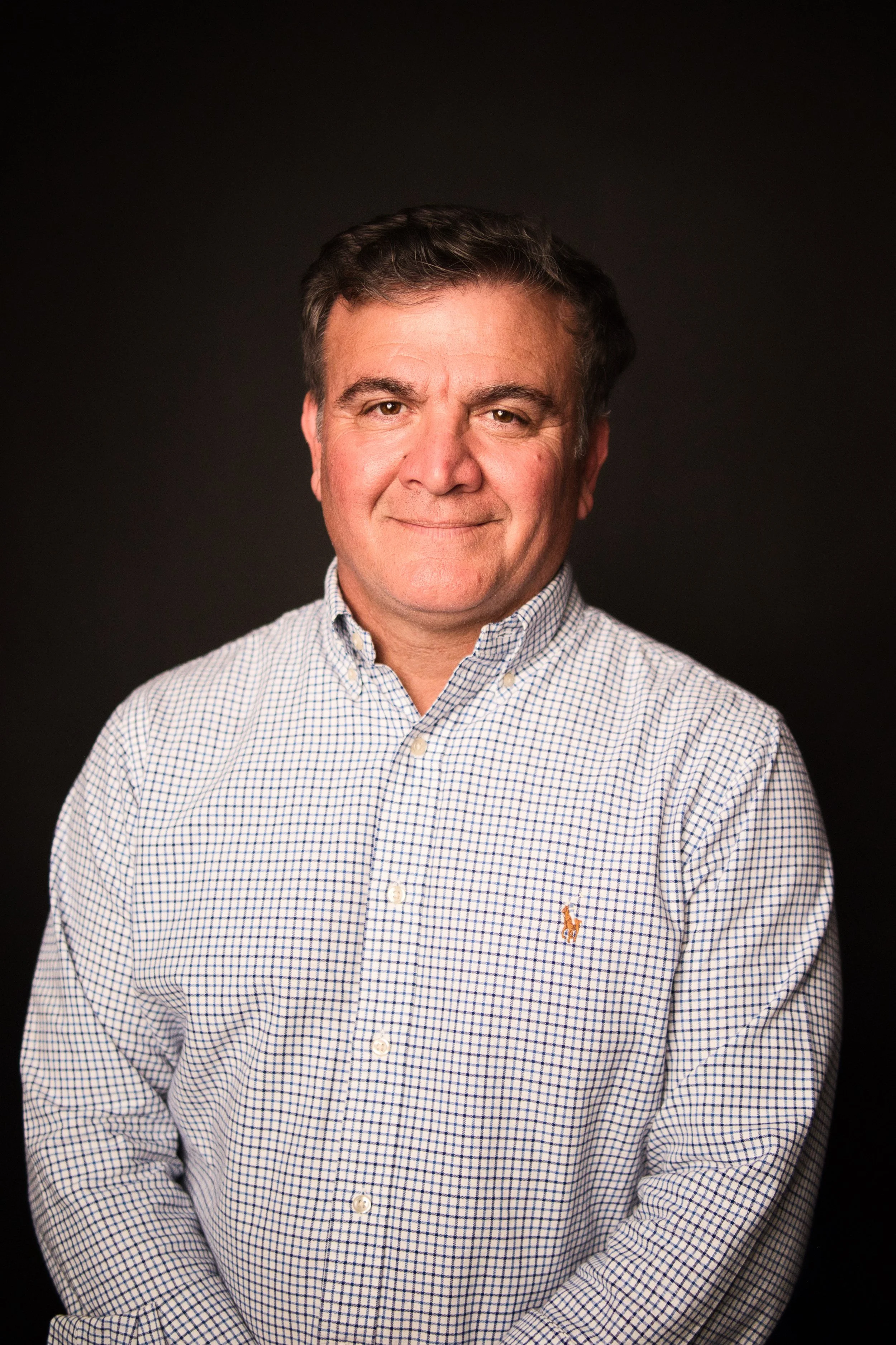 Portrait of a middle-aged man with short dark hair, wearing a checkered button-up shirt, smiling slightly, against a dark background.