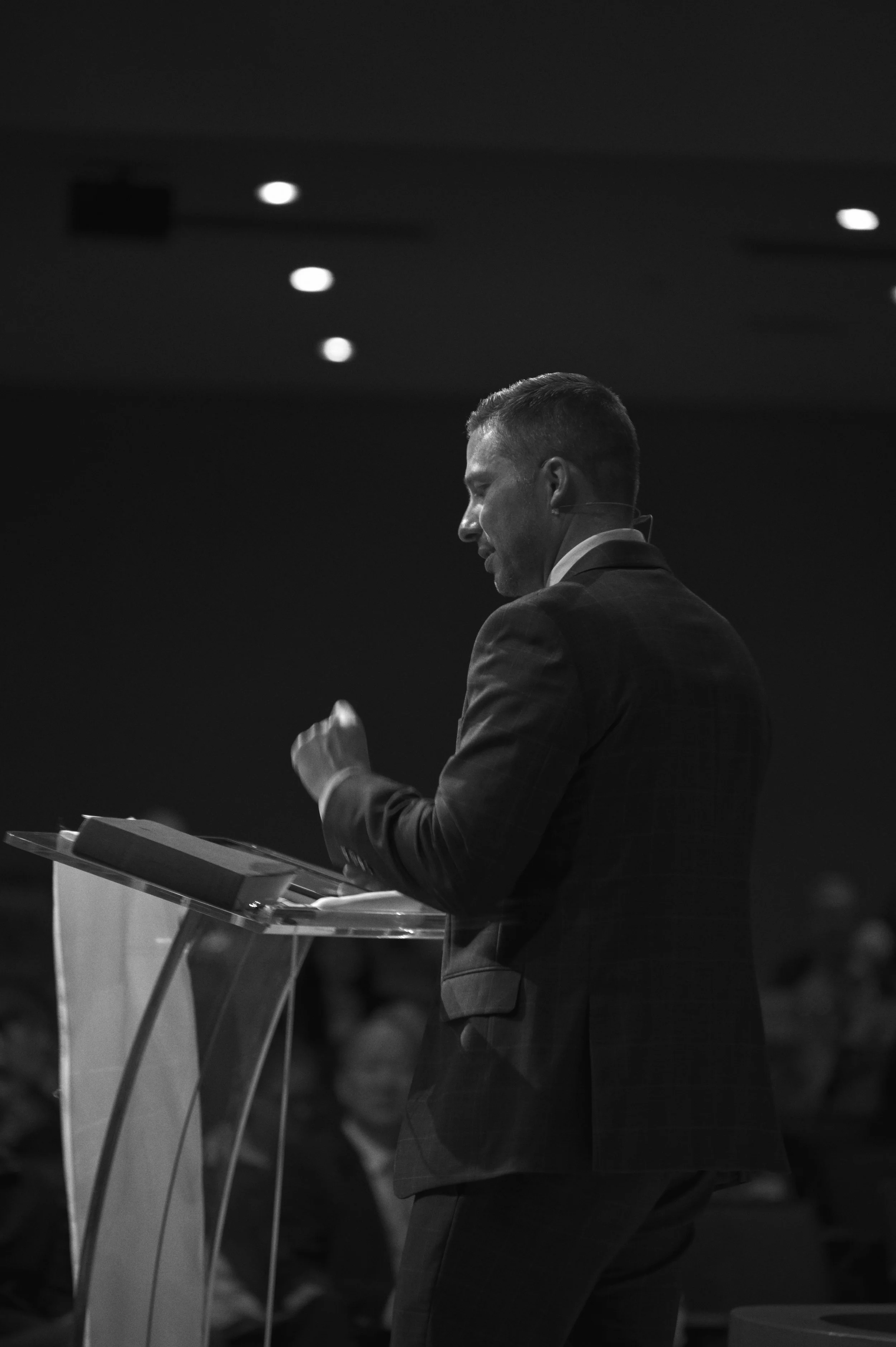 A man in a suit stands at a podium, speaking or presenting, with a lectern and papers in front of him, in a formal setting.