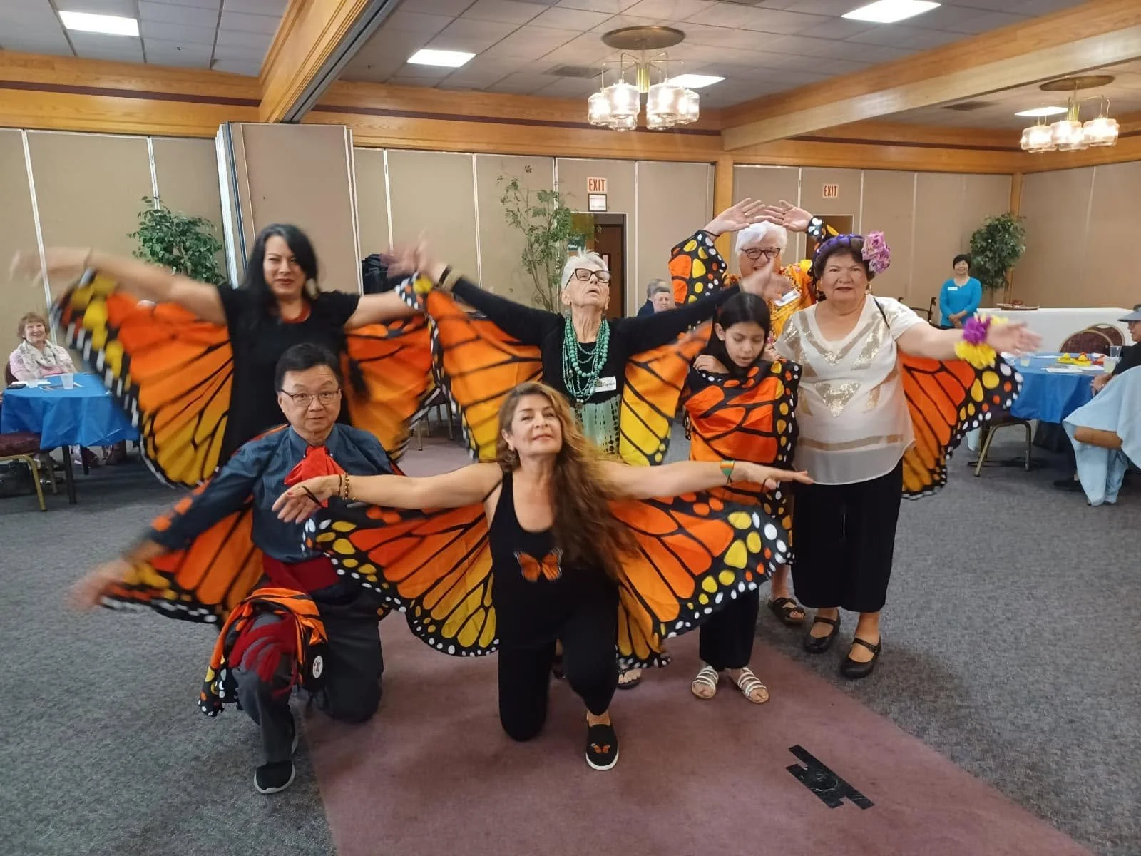 Group of people dressed as butterflies with orange and black wings, posing indoors at a social event or celebration, with tables and chairs in the background.