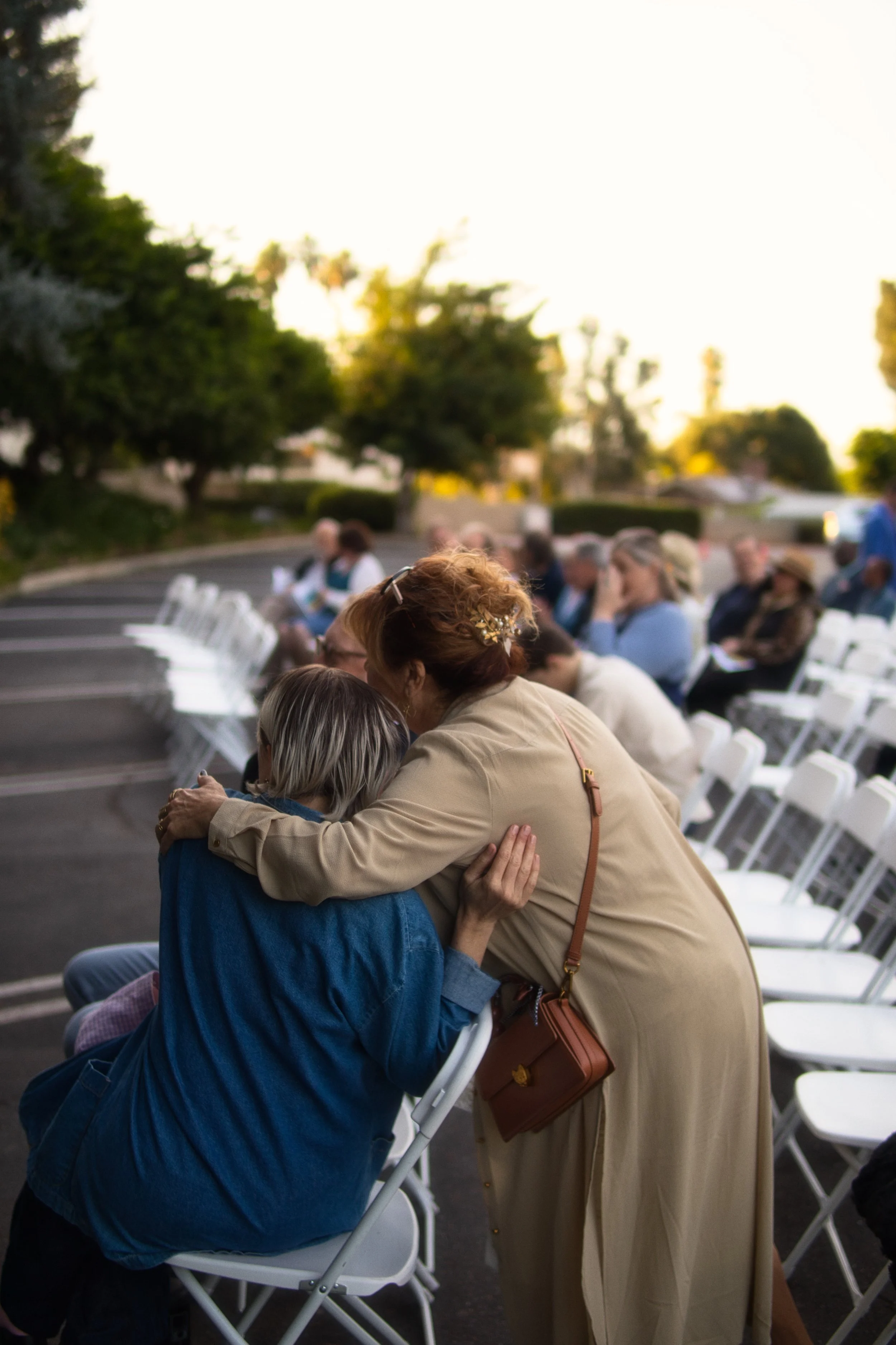 A woman embracing and comforting a person in a blue jacket amid sitting people outdoors at sunset.