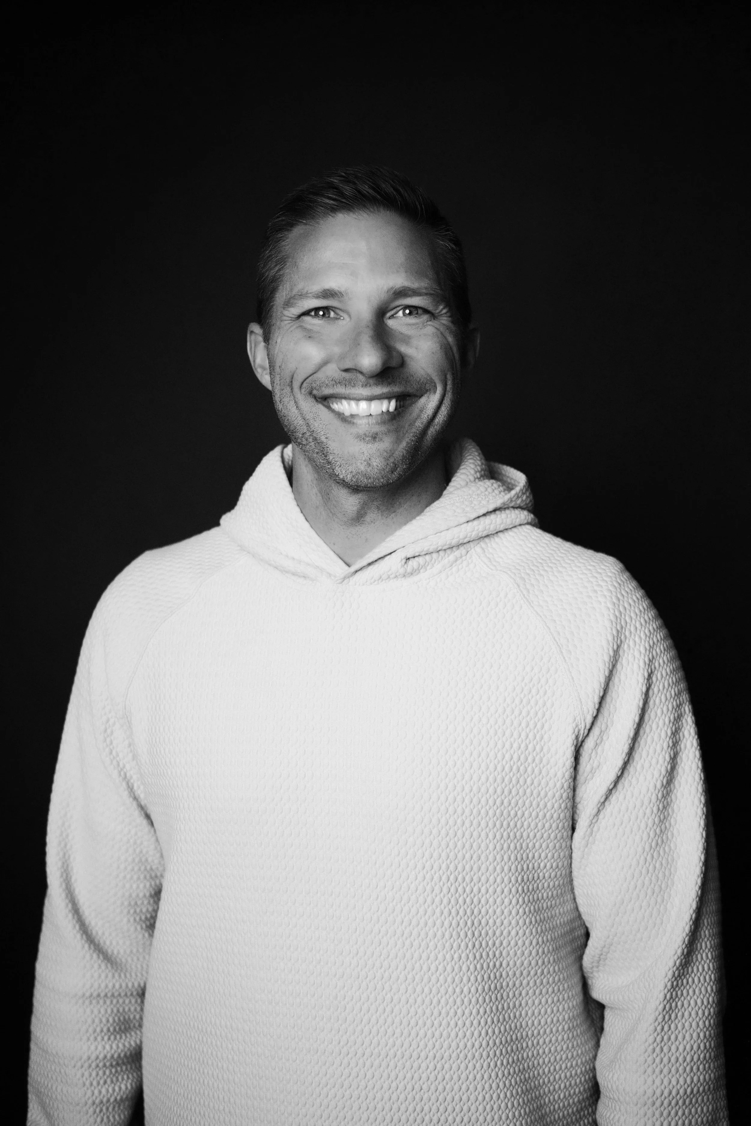 Black and white photo of a smiling man wearing a light textured hoodie, against a dark background.