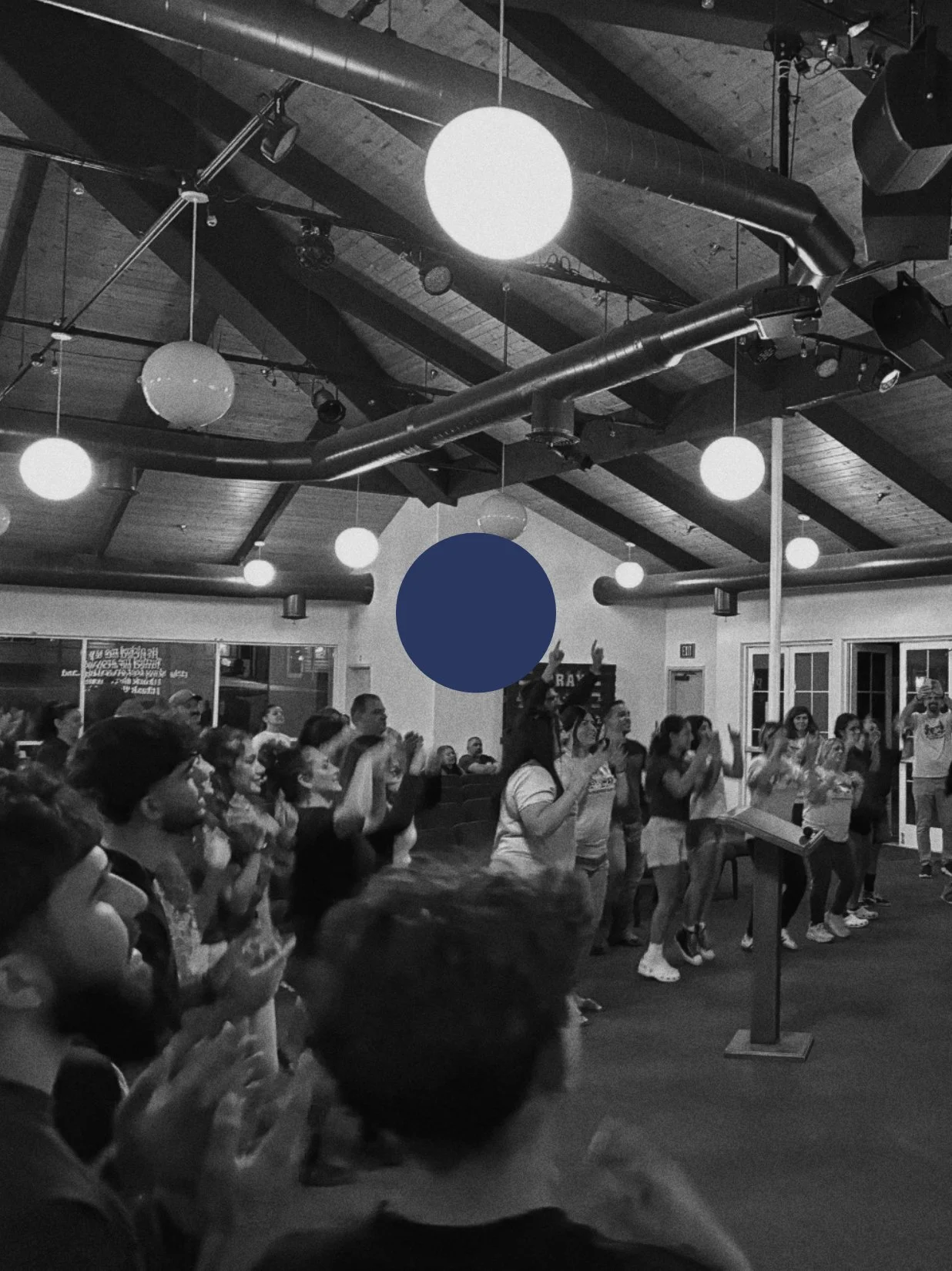 People are gathered in a room for a dance class or event, following the instructor who is at the front with arms raised. The room has a high wooden ceiling with large exposed beams, hanging round lights, and visible ductwork. The picture is in black and white.