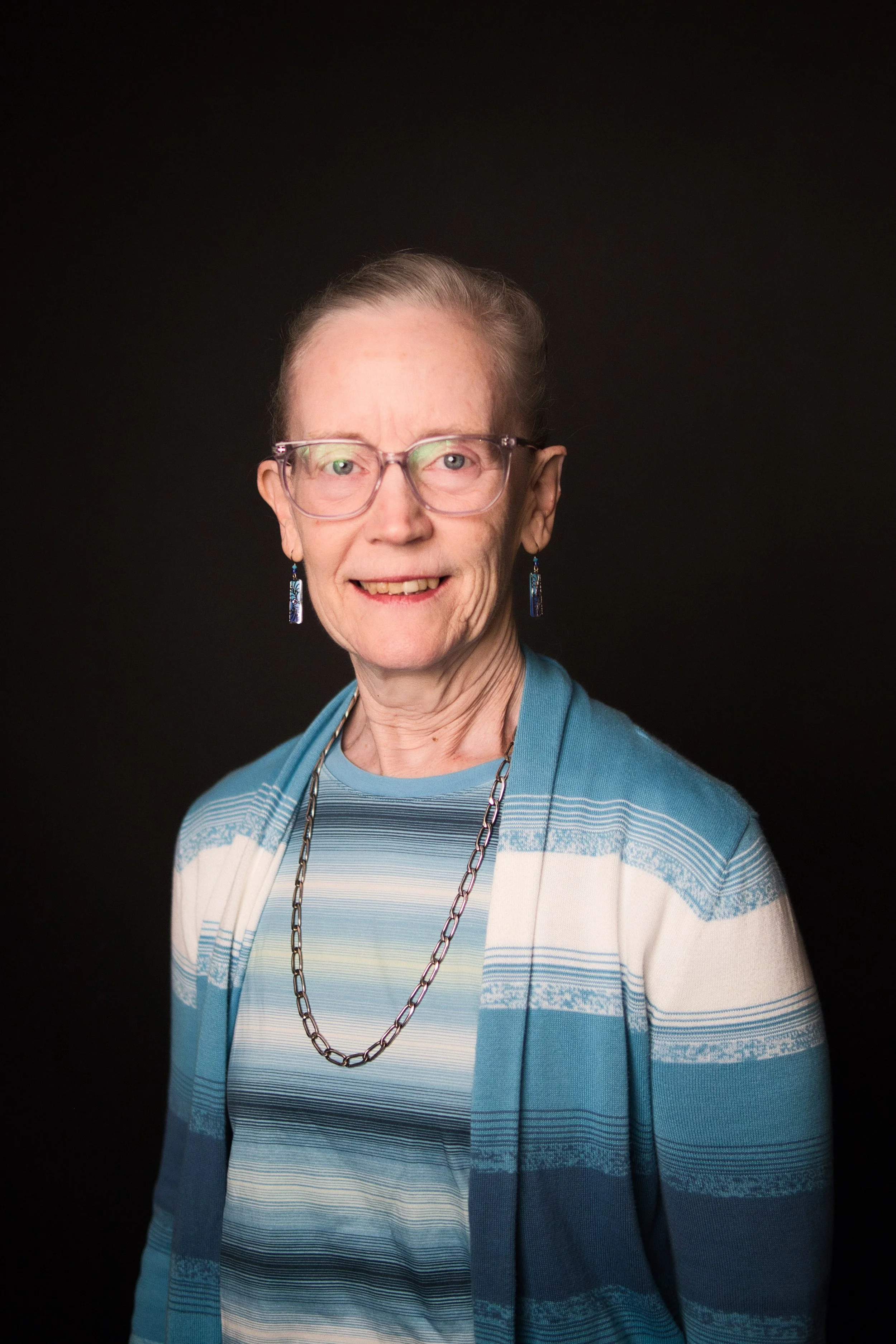 An older woman with short light brown hair wearing glasses, earrings, a silver chain necklace, a blue cardigan, and a striped shirt, smiling against a black background.
