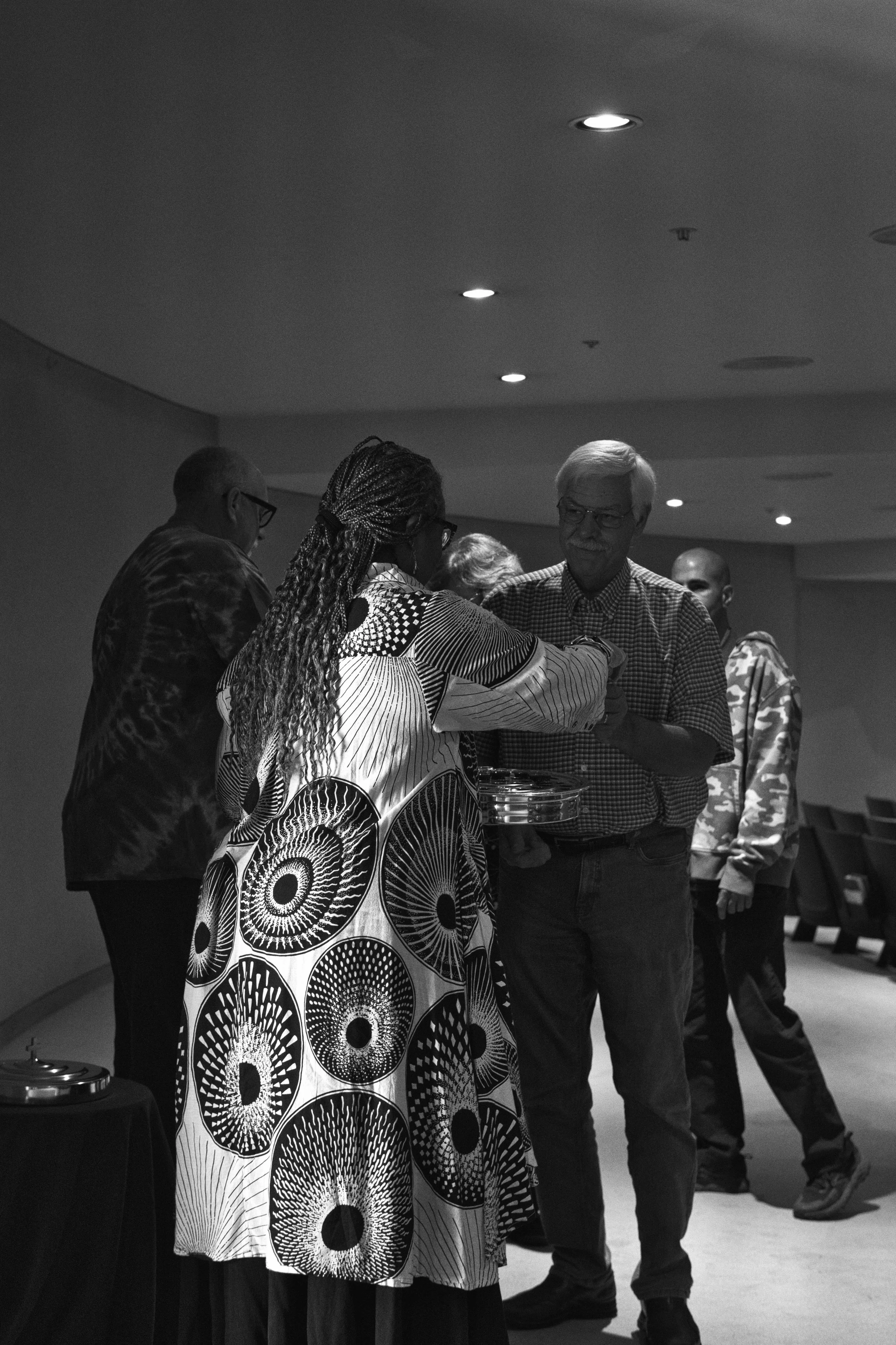 A black and white photo of a group of people in a discussion or greeting, with some standing around a table in an indoor setting with ceiling lights.