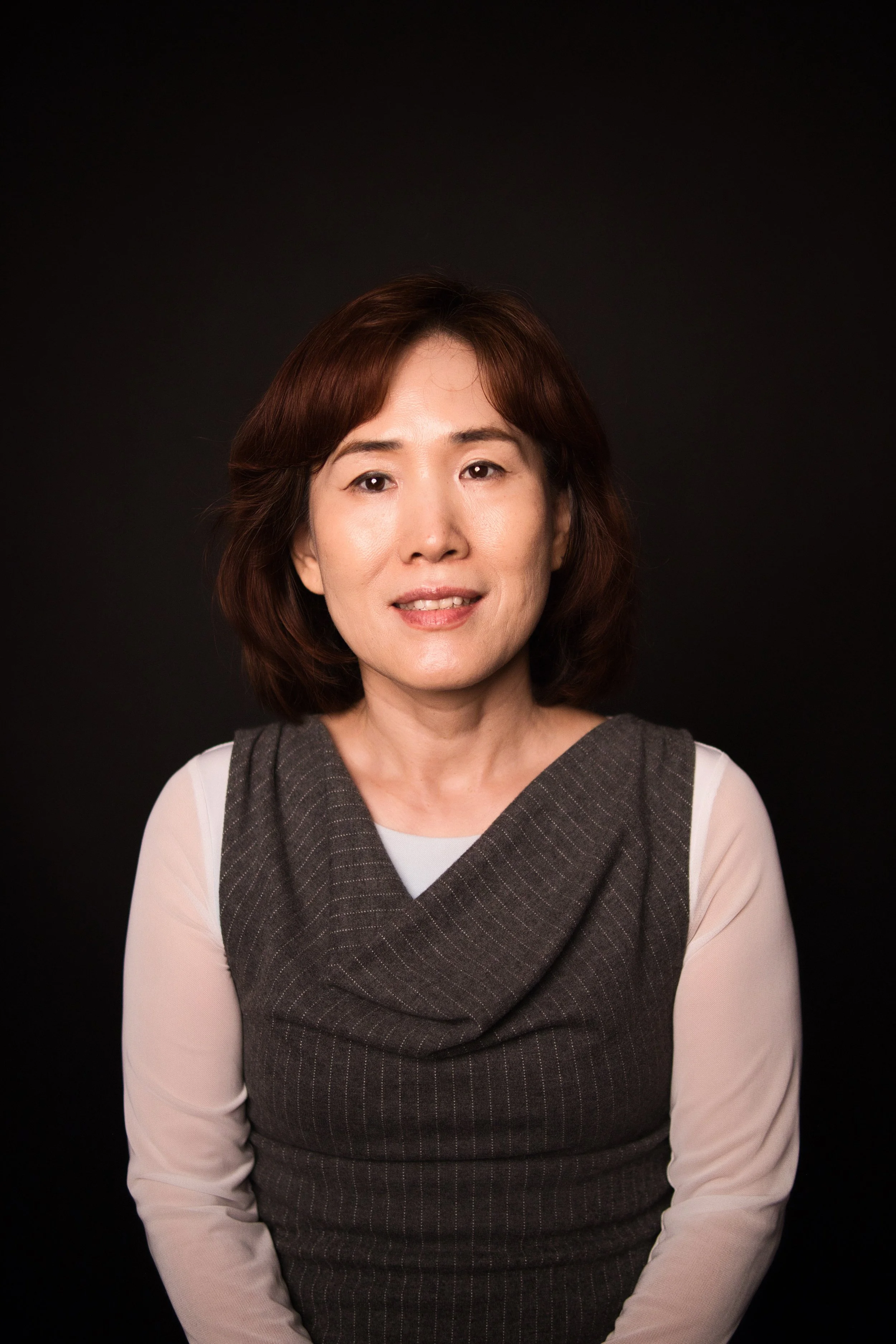 A woman with shoulder-length brown hair, wearing a grey sleeveless top over a white long-sleeve shirt, against a black background.