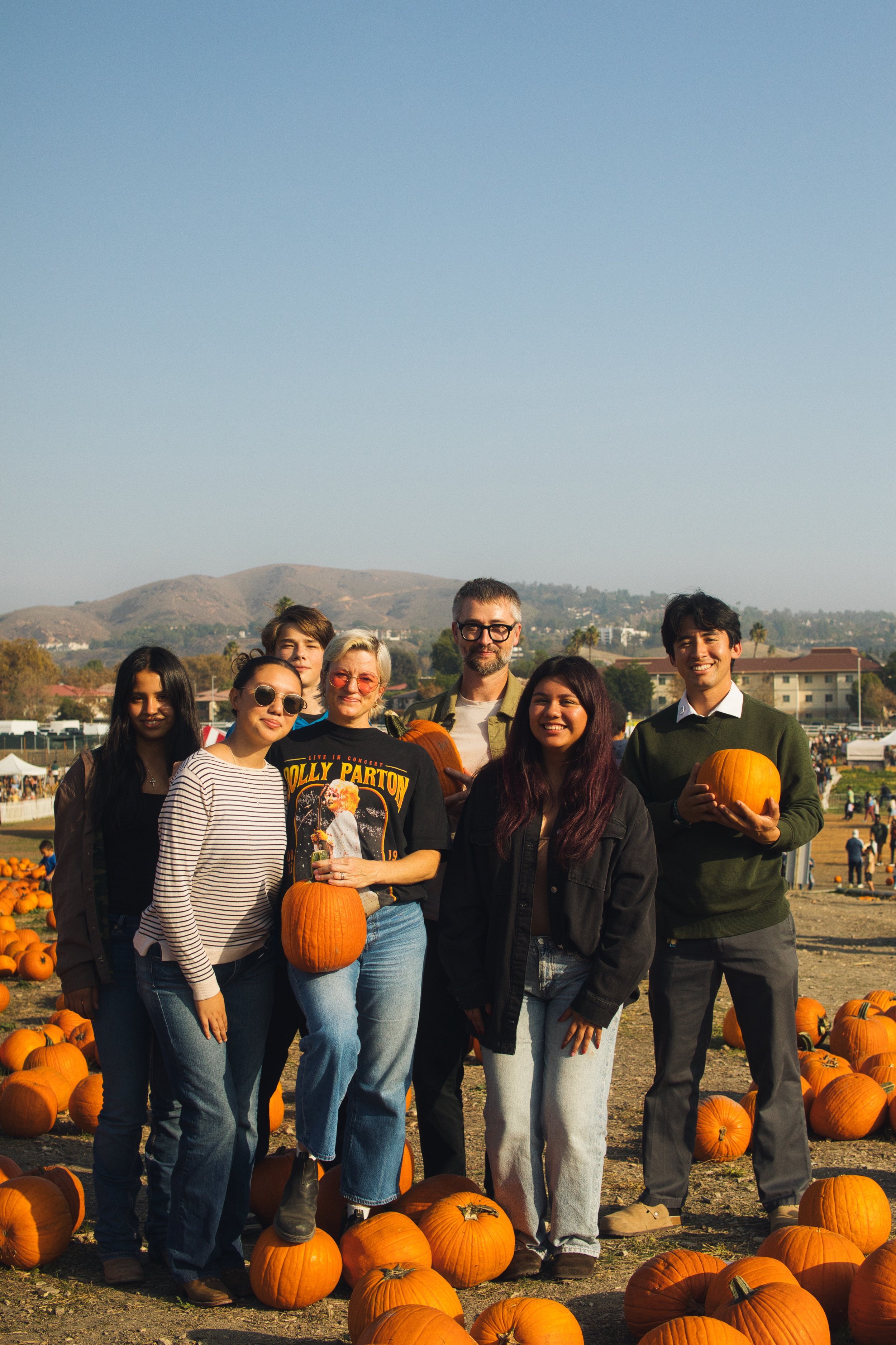 A group of people posing with pumpkins at a pumpkin patch during daytime with mountains in the background.