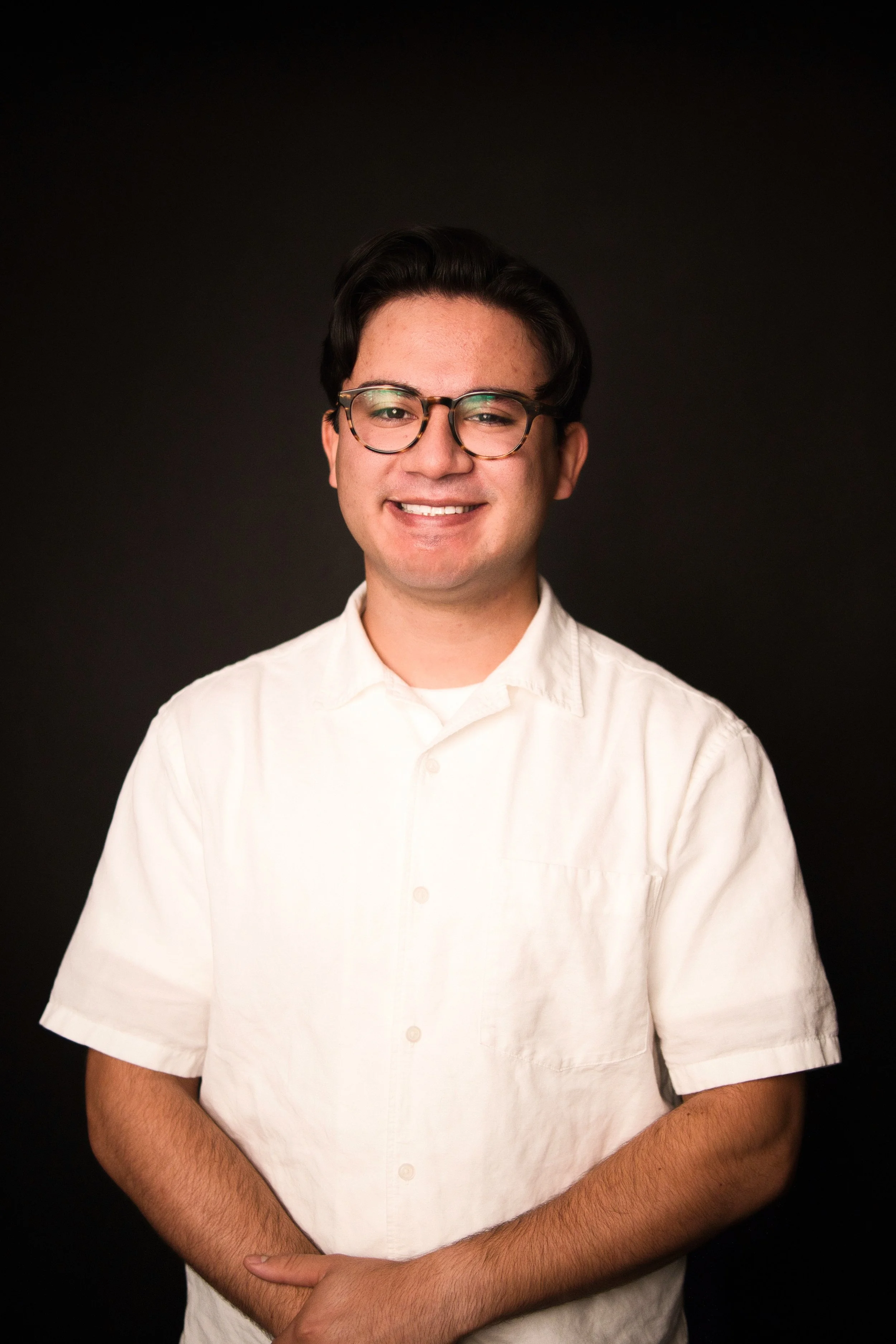A young man with dark hair, glasses, and a light complexion smiling, wearing a white short-sleeved shirt, against a plain dark background.