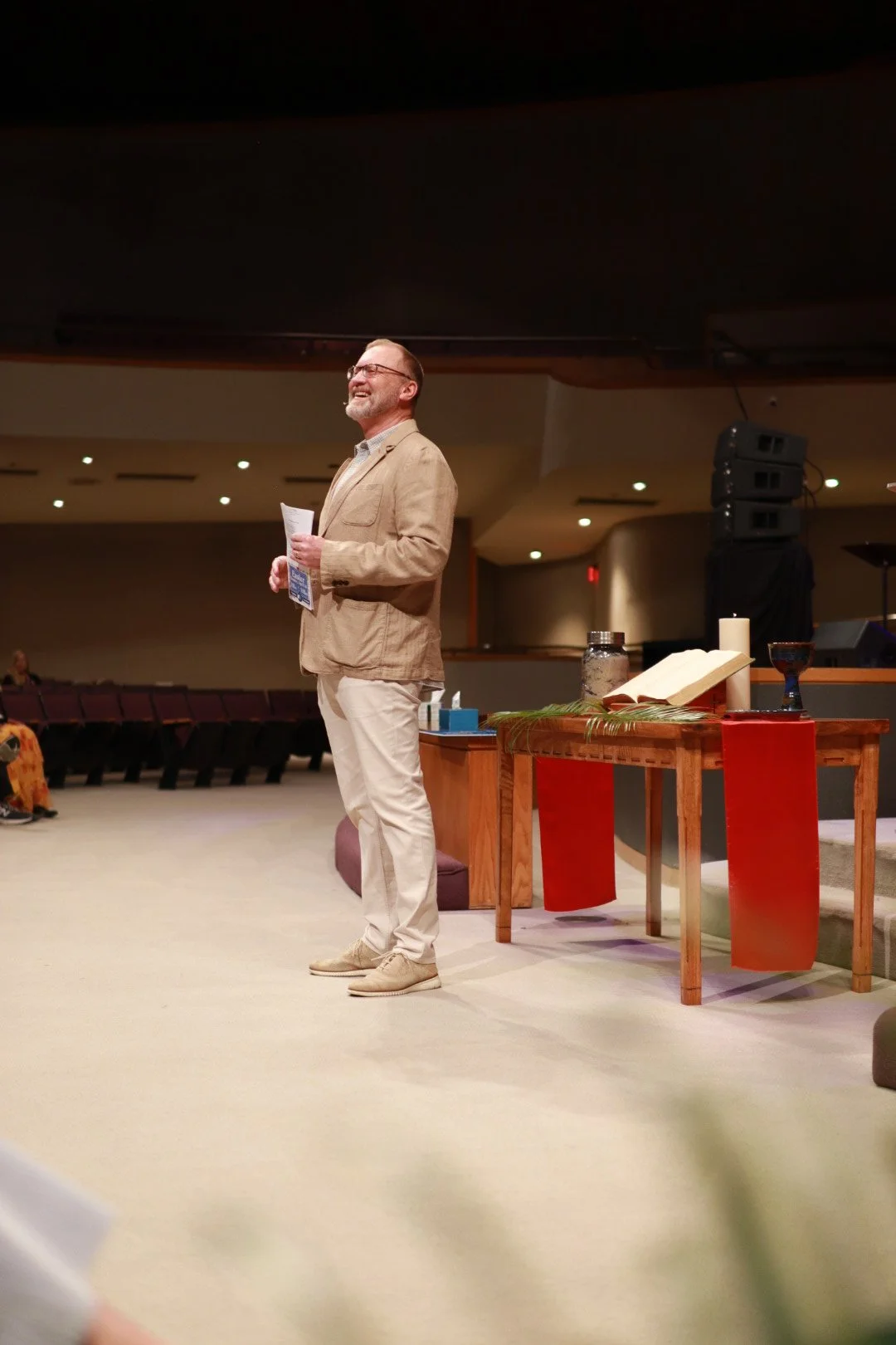 A man standing on stage in a beige blazer and white pants, holding papers, smiling. Behind him is a table with a candle, a book, and other items in a church or auditorium.