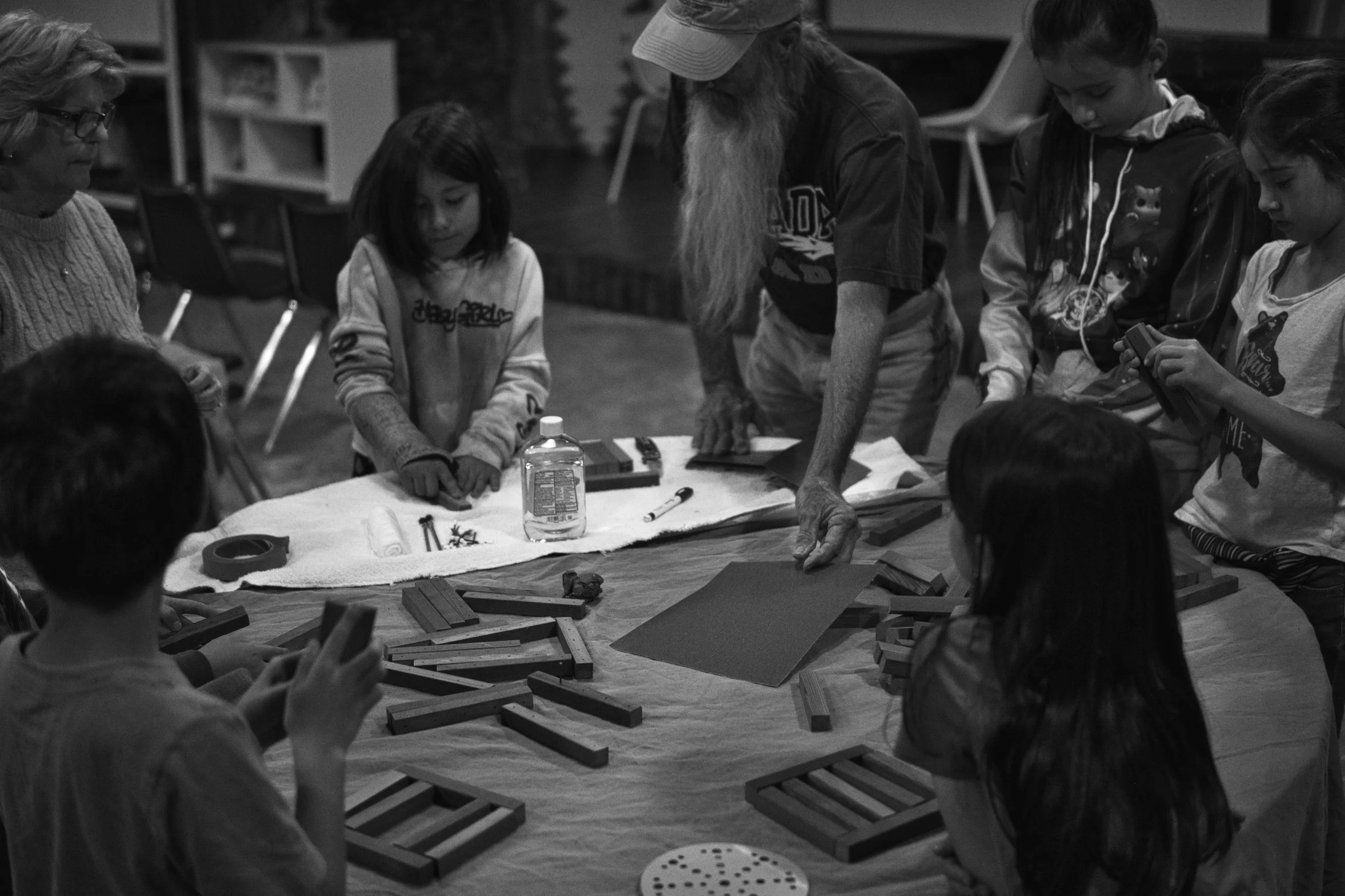 A group of children and an elderly man with a long beard gathered around a table, playing a tabletop game with wooden pieces, in a room with chairs and shelves in the background.