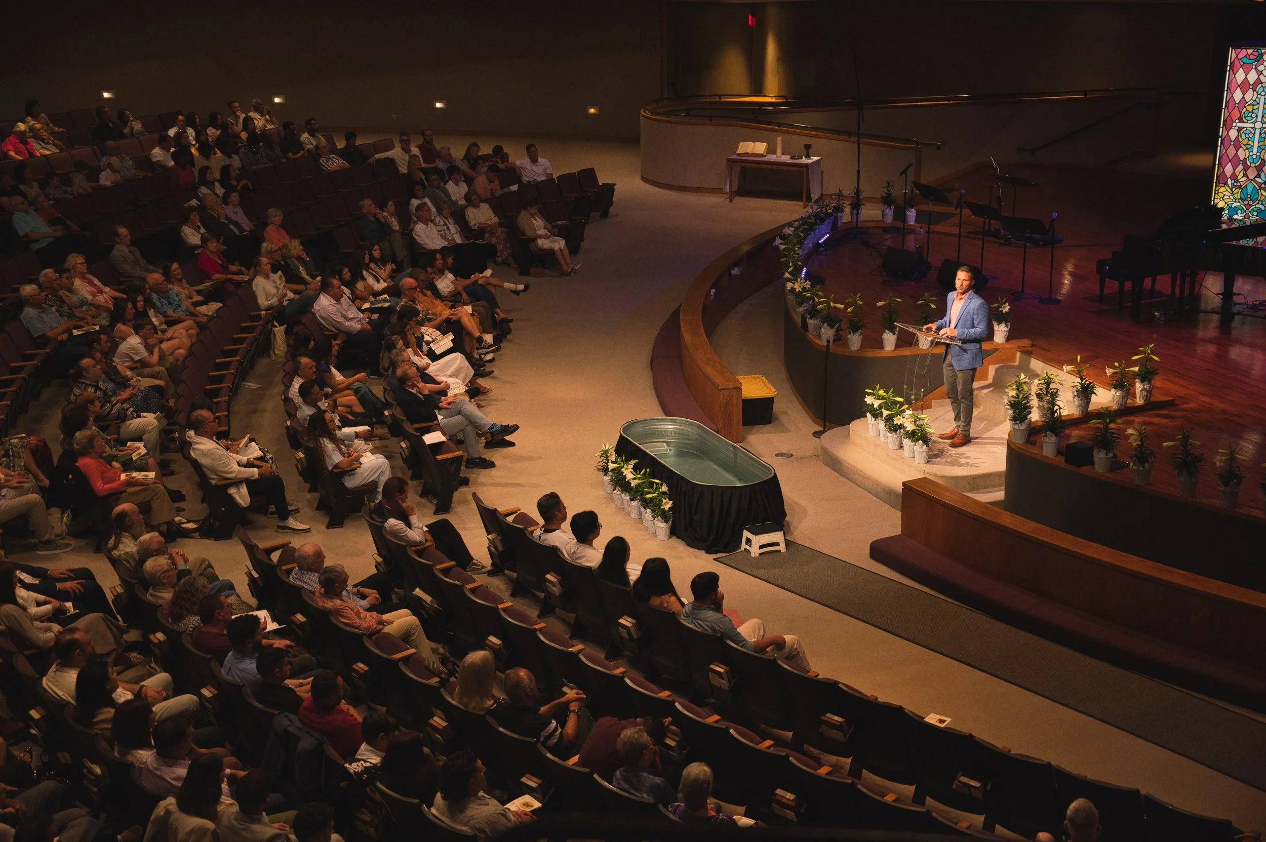 A speaker in a blue blazer is standing at a podium on a stage, addressing an audience seated in an auditorium. The stage is decorated with potted plants, and a stained glass window is visible on the right side.