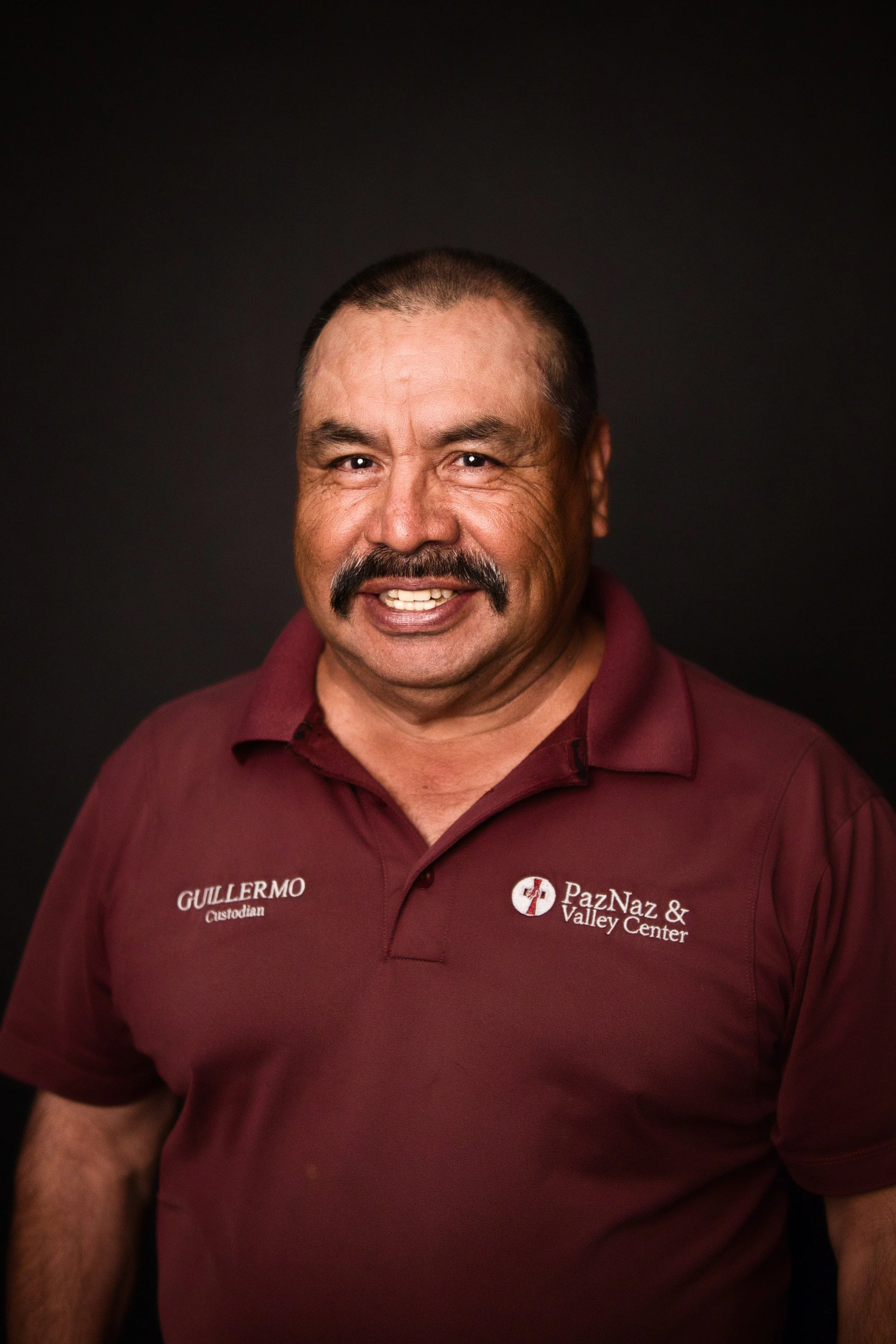 A man wearing a maroon polo shirt with 'Gullermo' and 'PazNaz & Valley Center' embroidered on it, smiling against a black background.