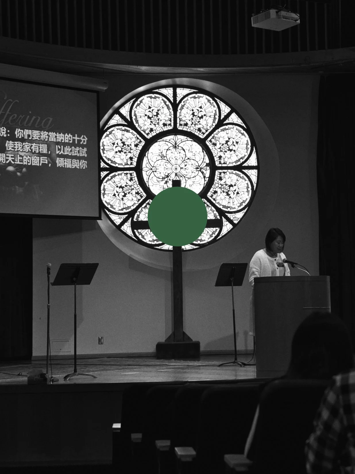 A woman is standing at a podium giving a presentation in a large room. Behind her, there is a large, ornate circular stained glass window. To the left, a projection screen displays text in Chinese. There are two microphone stands on either side of the podium, and the room appears to be set up for a formal event or lecture.