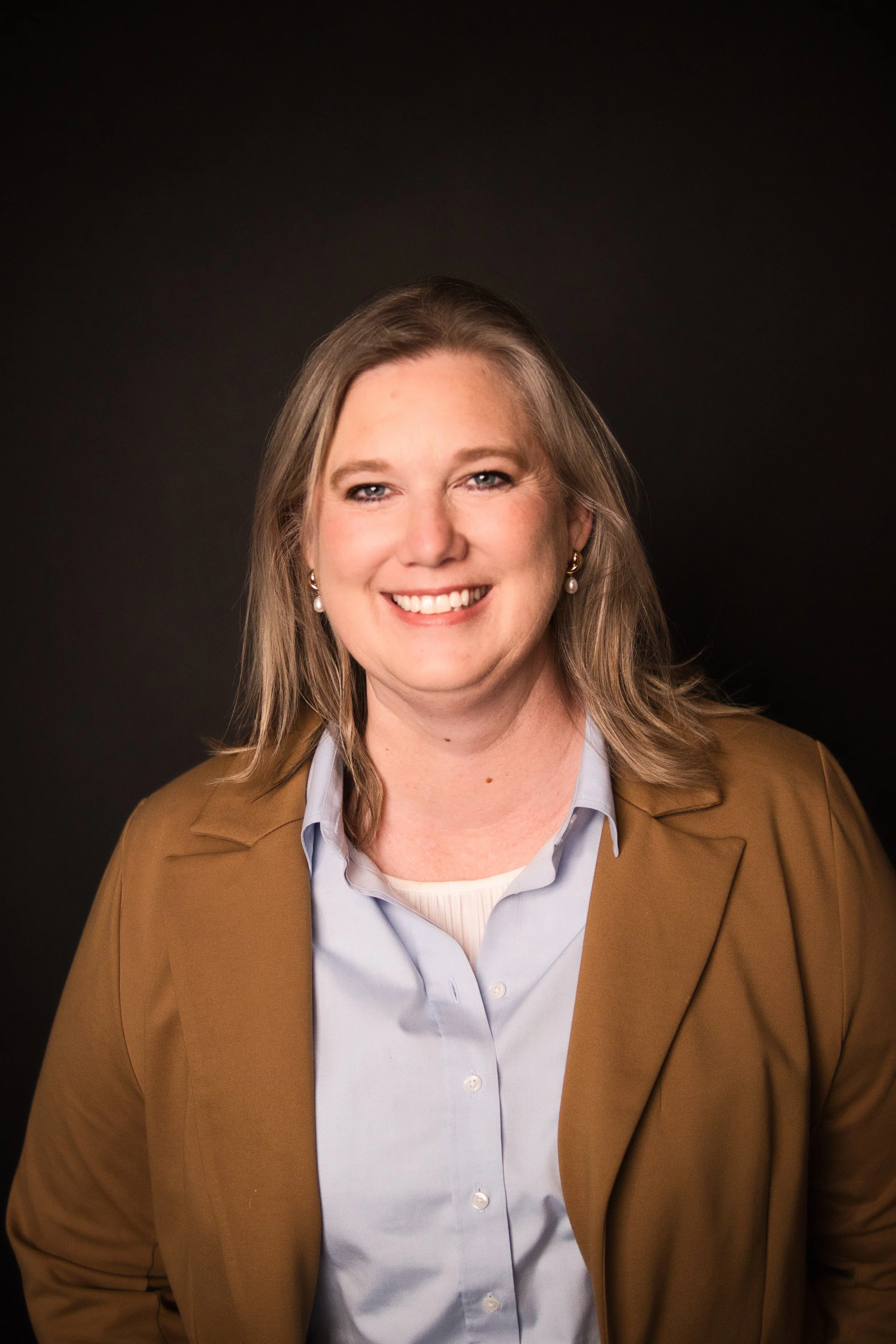 A smiling woman with blonde hair, wearing pearl earrings, a light blue collared shirt, and a brown blazer, standing against a dark background.