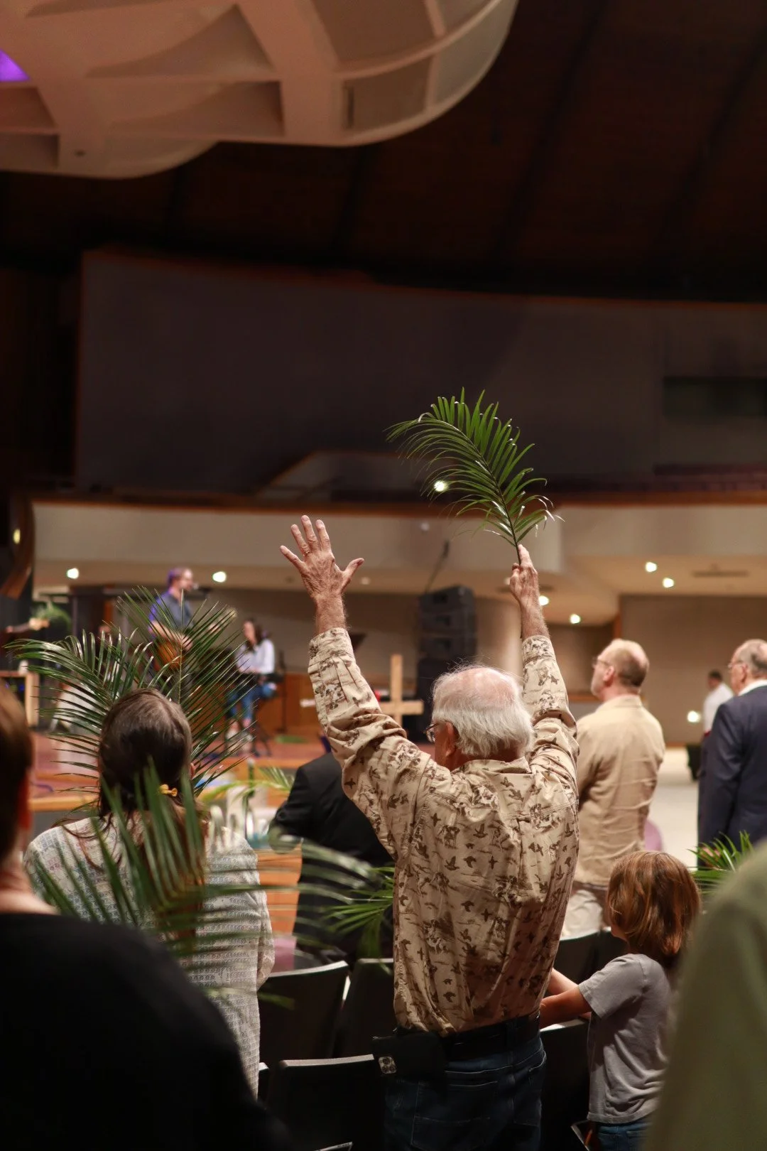 An elderly man in a camouflage shirt raising his hand with a palm branch in a crowded indoor setting, possibly a church or event hall, with other people and musicians on stage in the background.