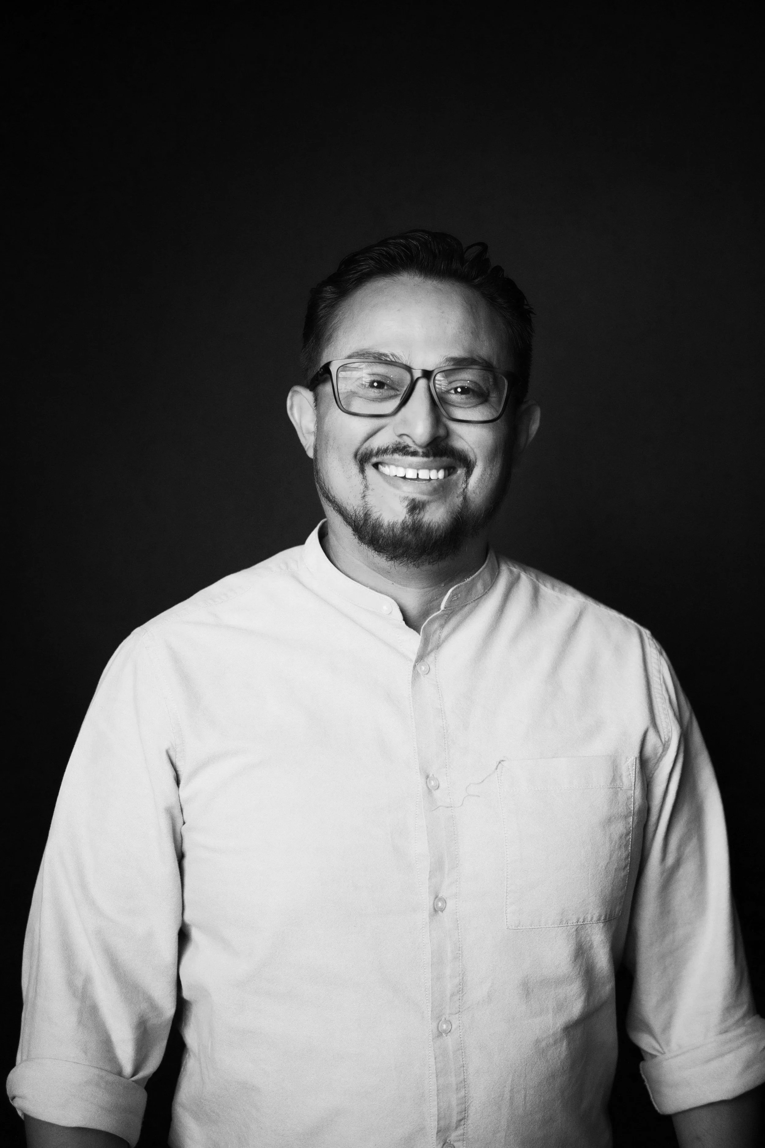 A black and white portrait of a man smiling, wearing glasses and a light-colored collared shirt, against a dark background.