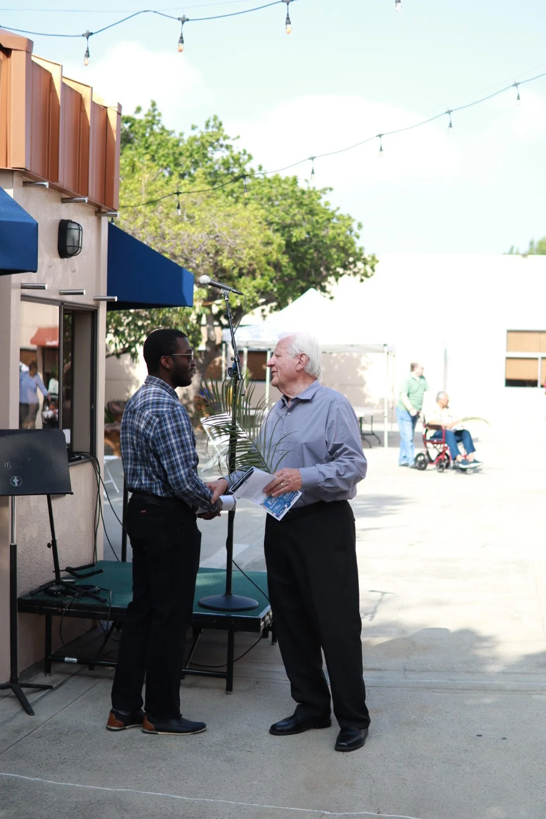 Two men standing and talking outdoors, one holding a leaf and a booklet, with a microphone stand nearby. In the background, people are sitting and walking in a sunny, open area with string lights overhead.
